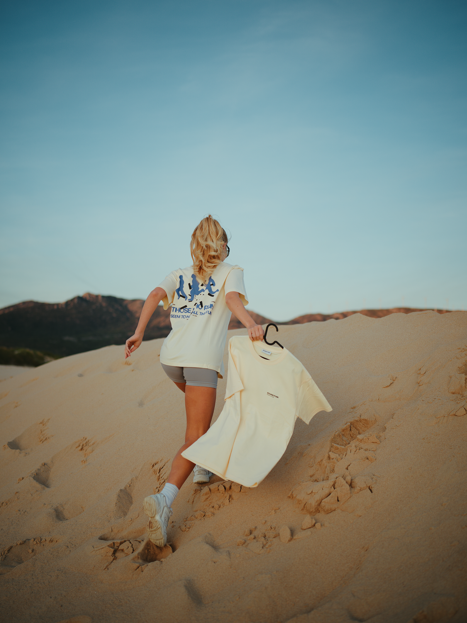 Person walking up sandy dunes holding a cream-colored t-shirt on a hanger, with mountains in the background and a clear blue sky.