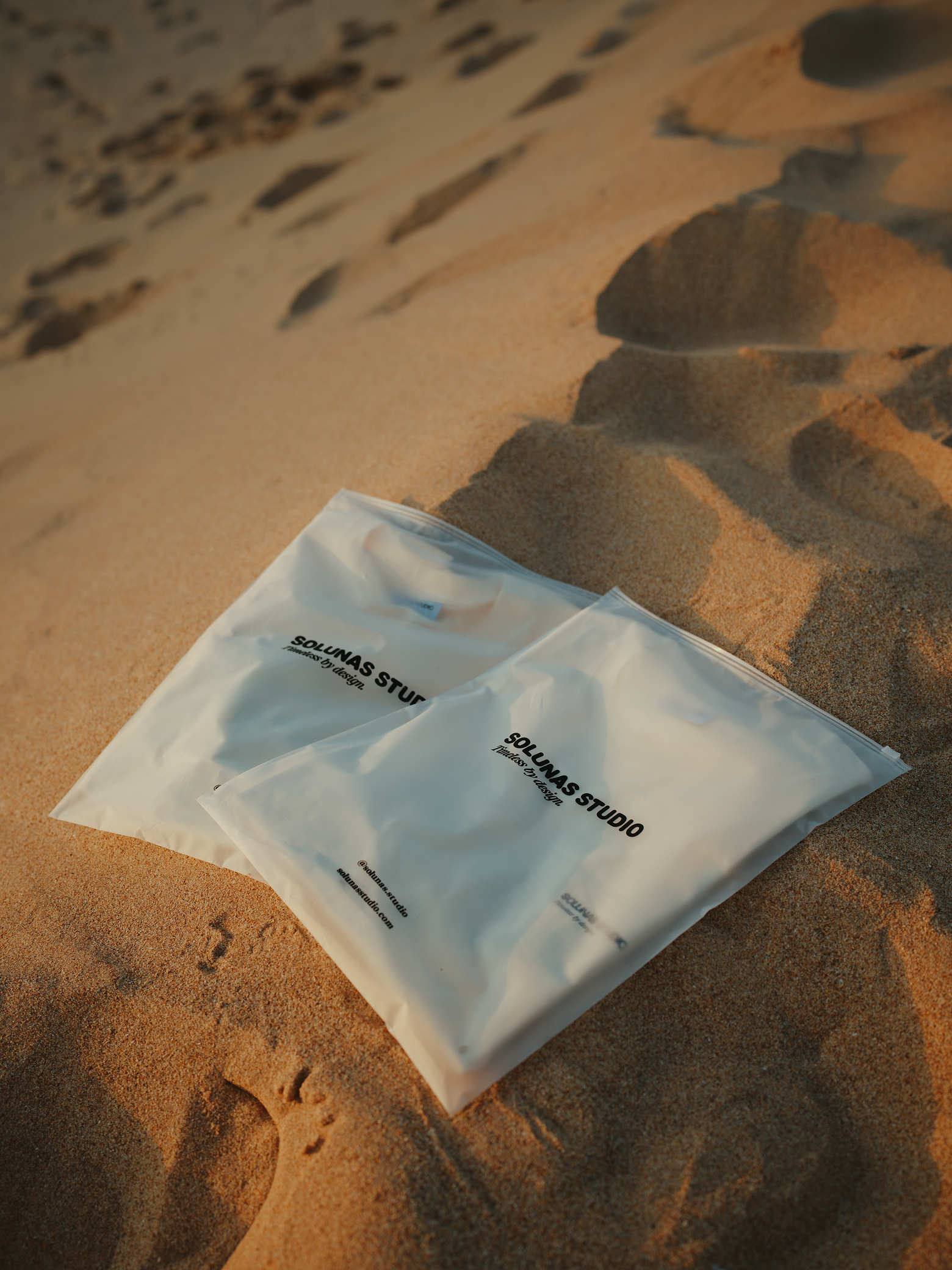 Two plastic bags with 'SOLUNAS STUDIO' logo lying on sandy surface, some rocks in the background.