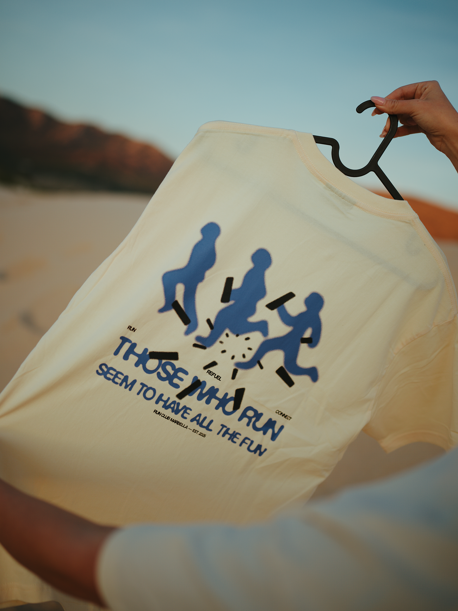A person holding a yellow t-shirt with a graphic of four people running and the text 'THOSE WHO RUN SEEM TO HAVE ALL THE FUN' printed on it, against a background of a blue sky and mountainous landscape.