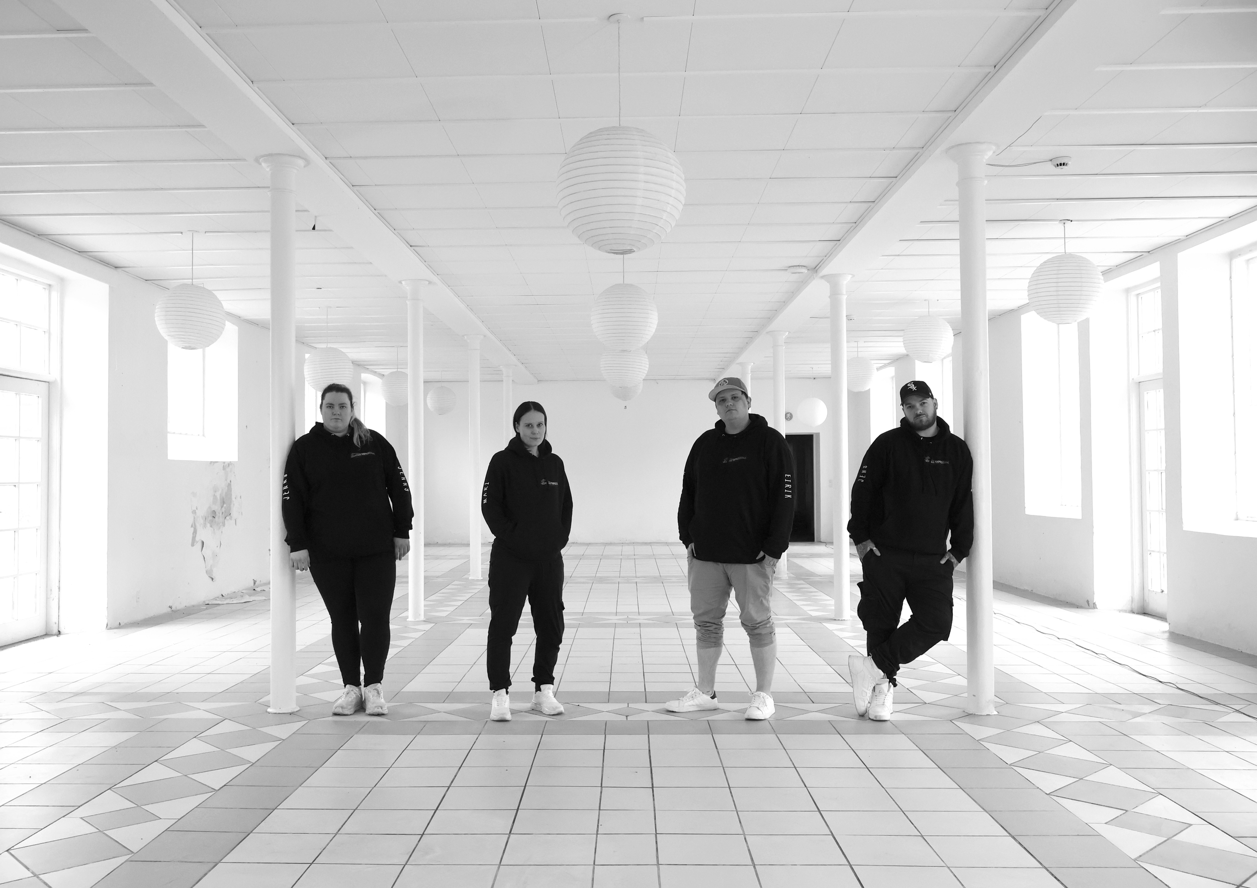 Four people standing in a spacious, empty room with white walls, large windows, tiled floor, and hanging paper lanterns, all wearing black hoodies and casual pants.