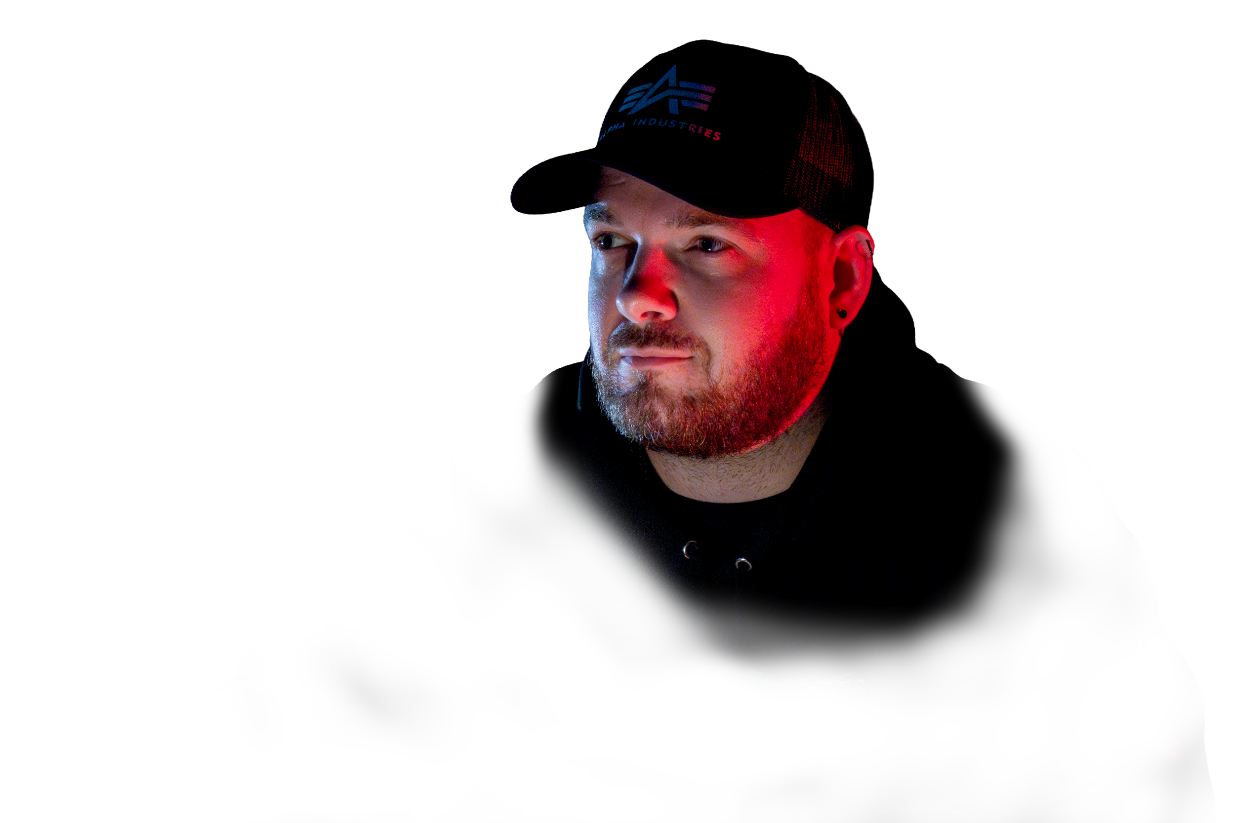 A man with a beard and earring sitting at a table, wearing a black cap and hoodie, illuminated by red and white lighting against a dark background.