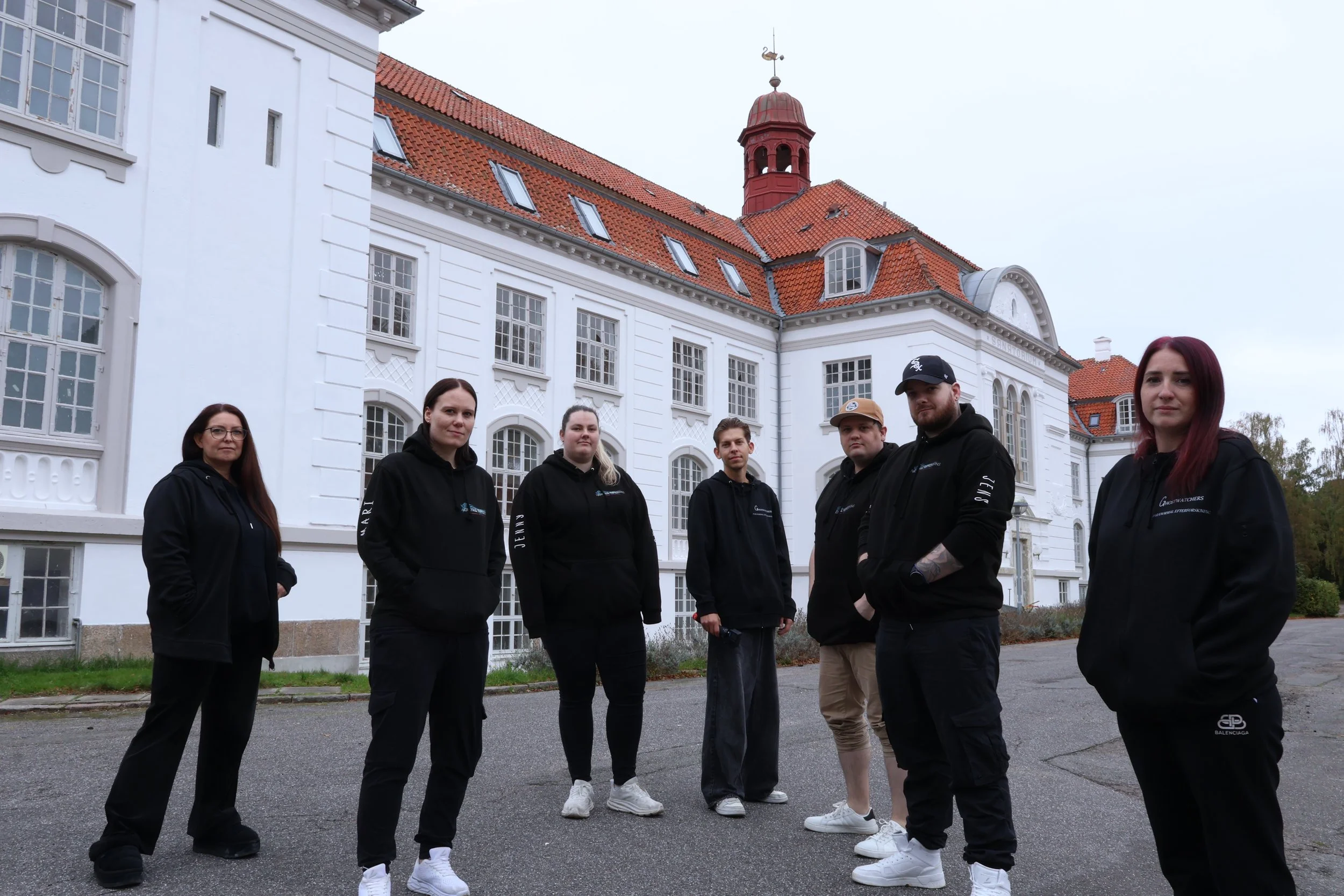 Group of seven people standing outside on a paved area in front of a large white building with red-tiled roof and multiple windows, some with dormers, under an overcast sky.