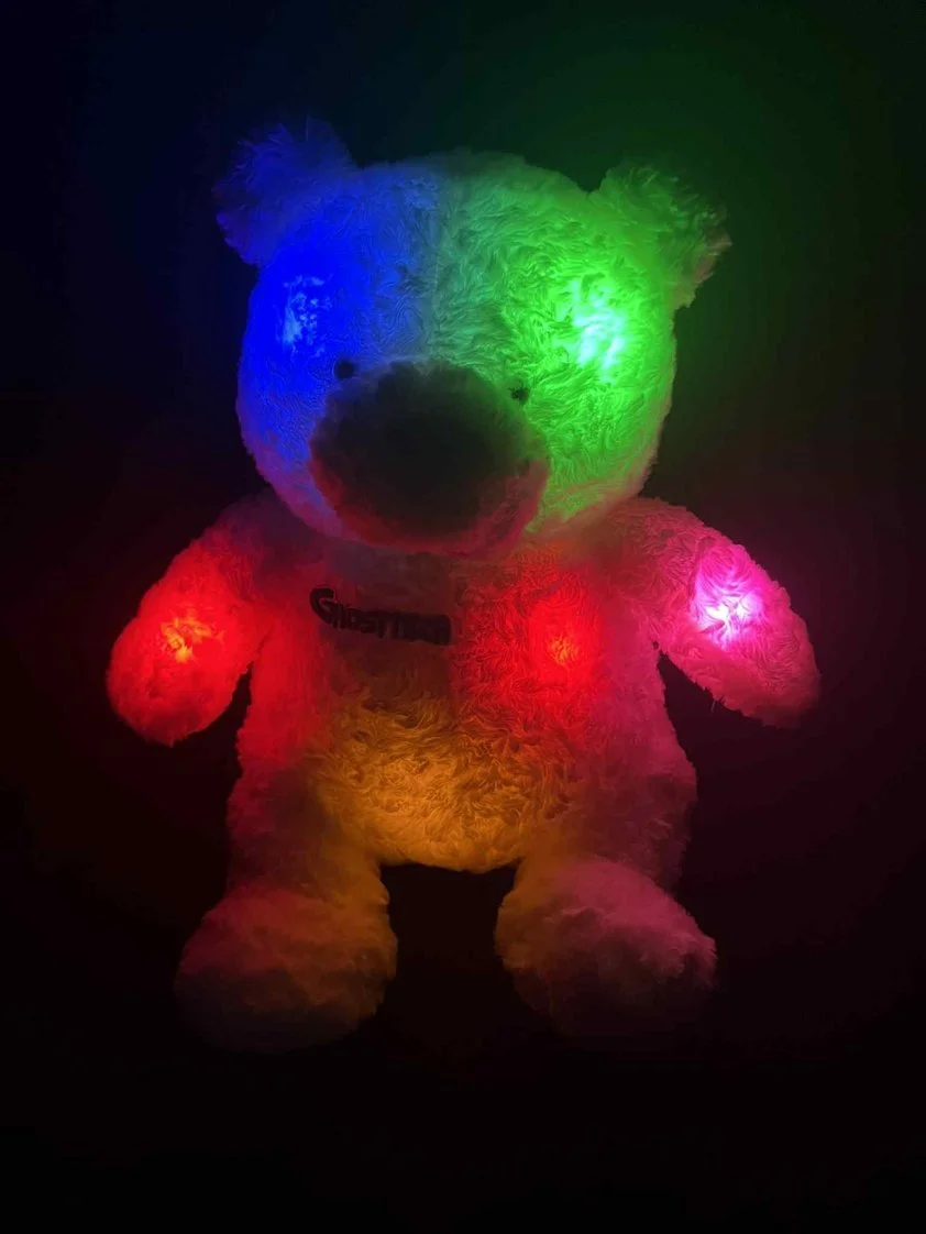 Plush teddy bear with multicolored LED lights illuminating its face and body, displayed in a dark setting.