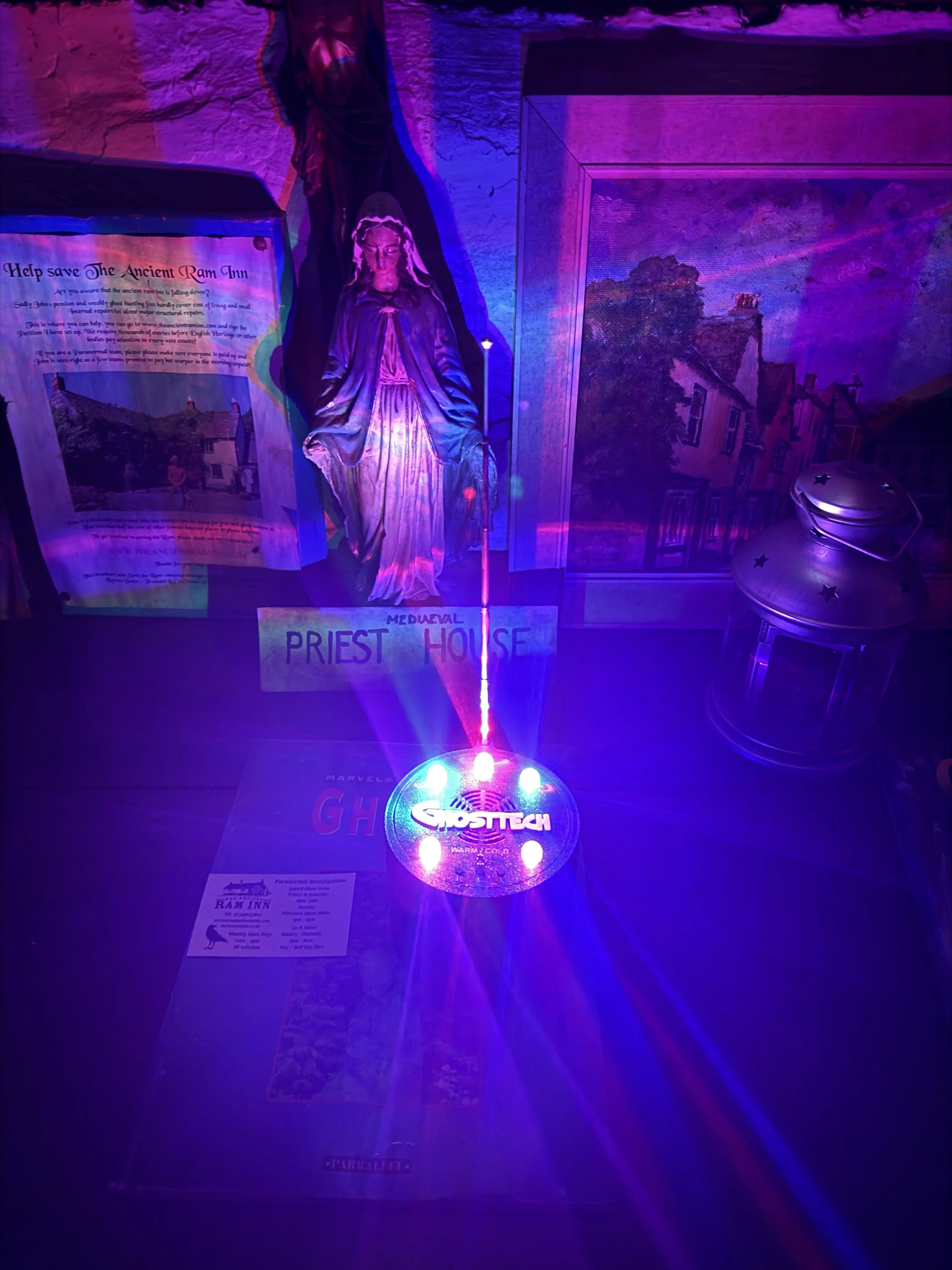 A display with a statue of a priest or religious figure, framed pictures, and a colorful GhostTech light with purple and blue beams.