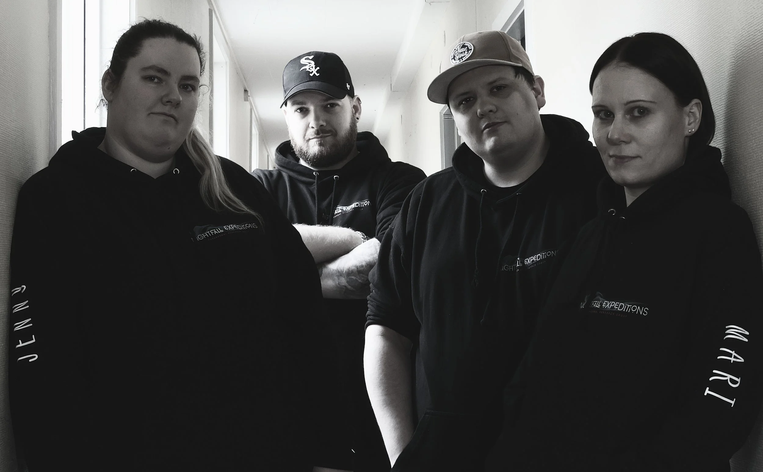 Four young adults wearing black hoodies standing in a hallway, posing for the camera with neutral expressions.
