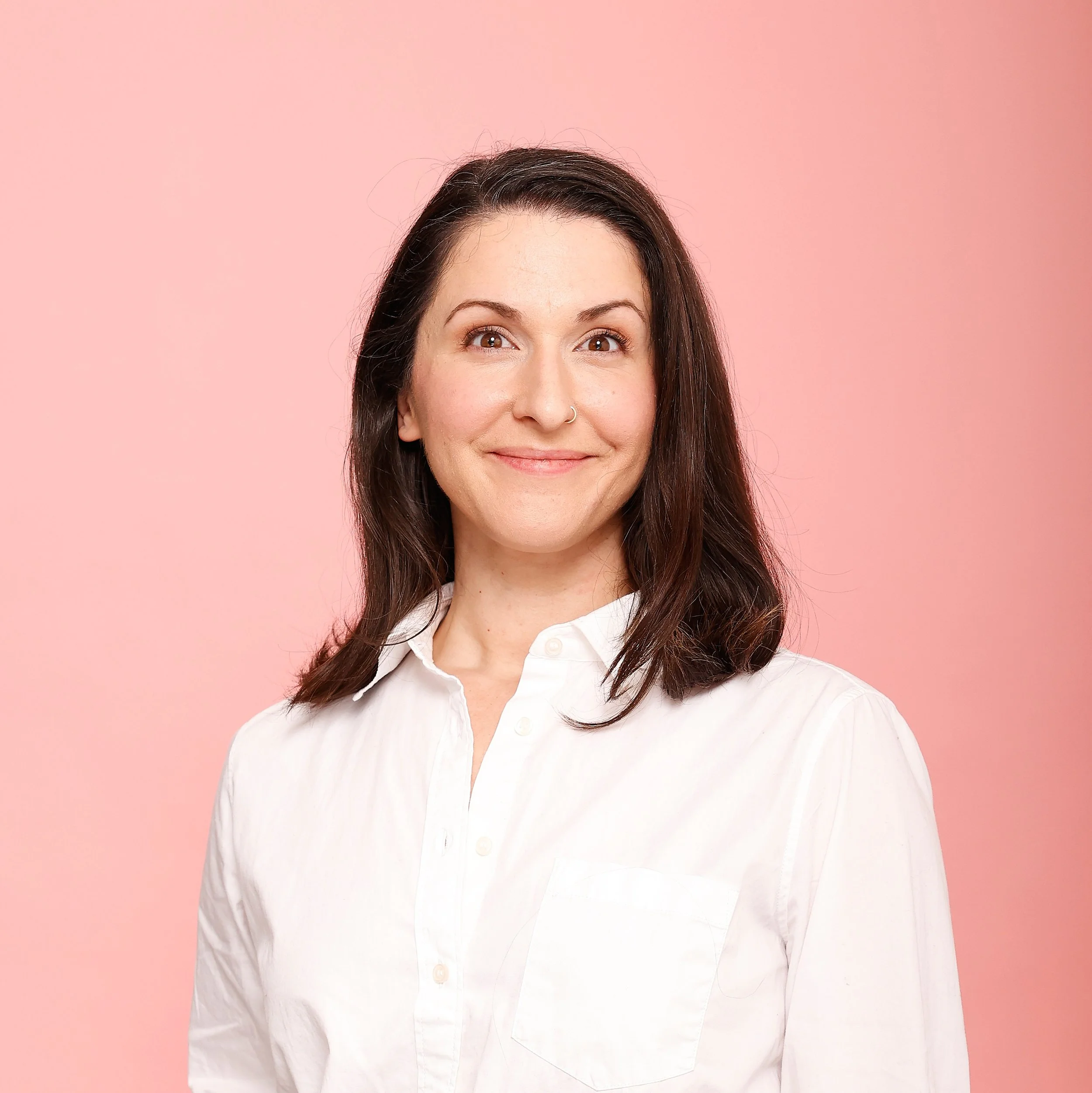 Headshot of Ashley Thimot. Ashley is in front of a pink background and wears a white shirt. She is smiling at the camera and has brown hair.