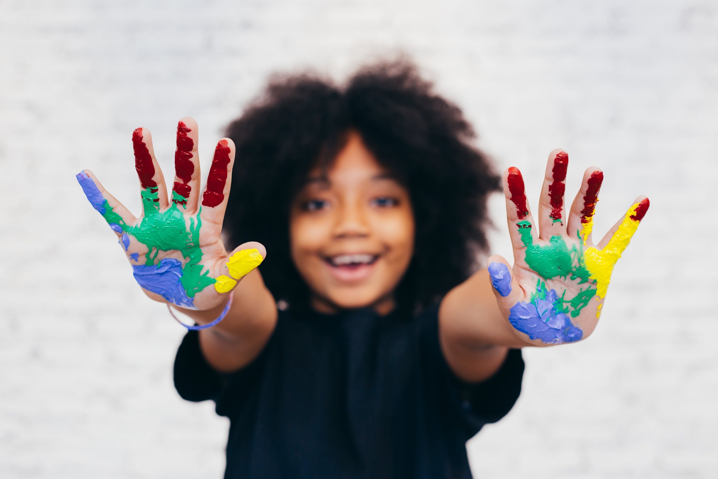 Picture of a young child smiling with their hands extended forward covered in blue, green, red, and yellow paint.