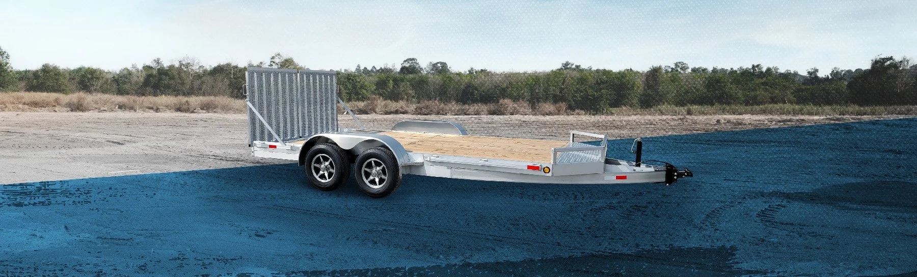 Empty flatbed utility trailer on a dirt surface with a patch of blue material underneath, against a background of trees and a cloudy sky.