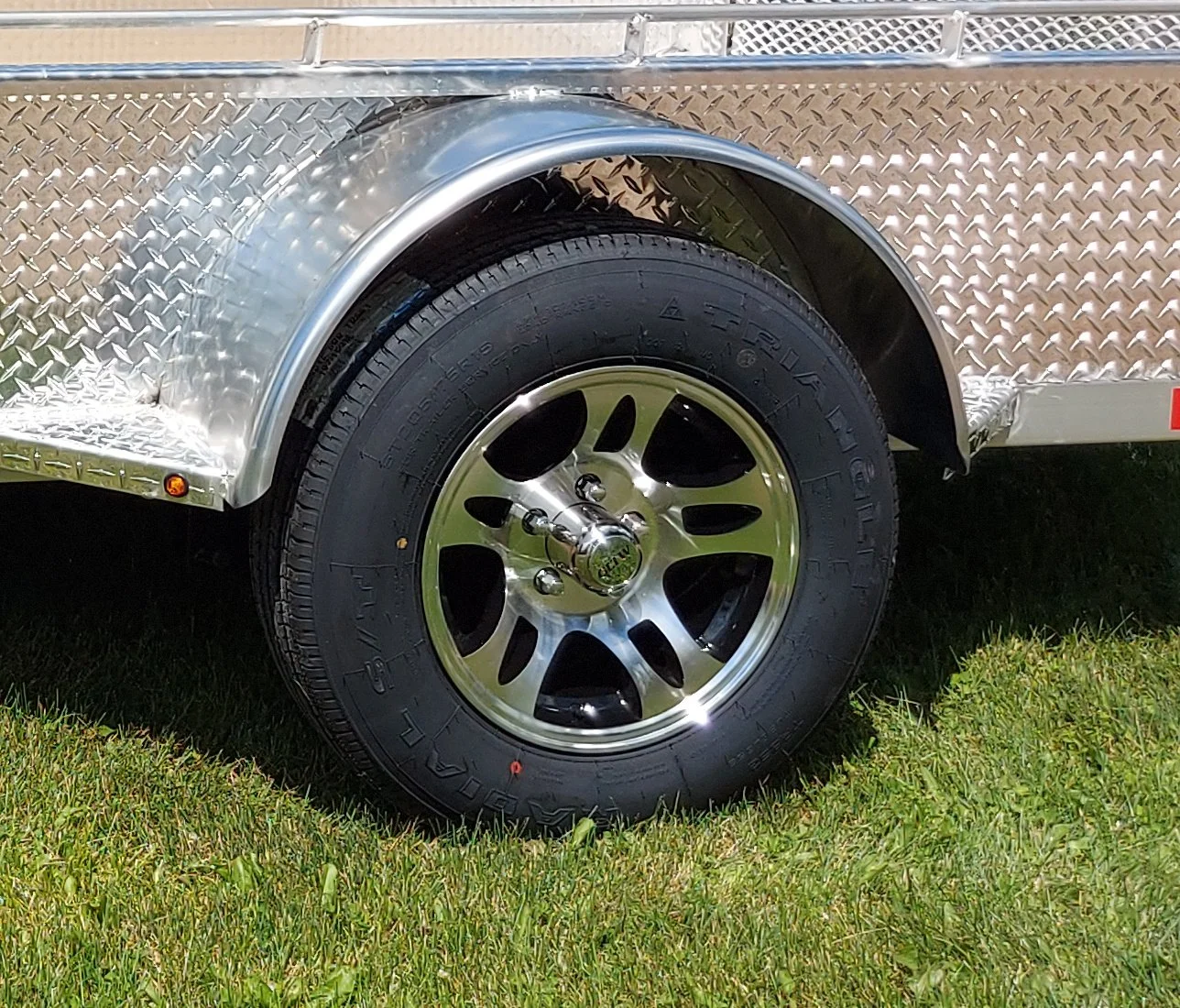 A close-up of a black tire on a shiny aluminum wheel, mounted on a metal trailer with diamond plate sides, resting on green grass.