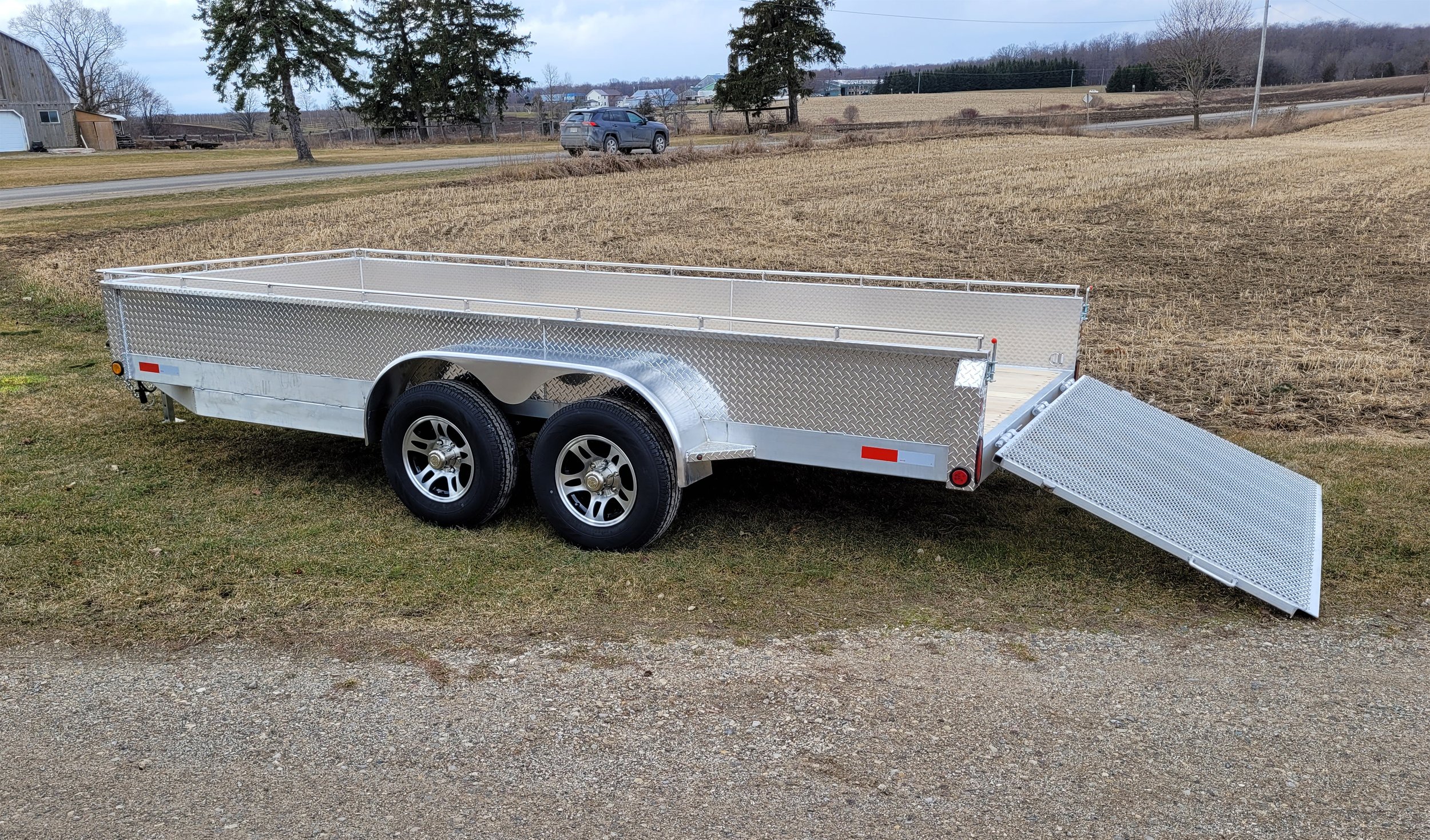 Silver utility trailer with dual axles on a grassy area, with a ramp lowered at the back, and a rural landscape in the background.