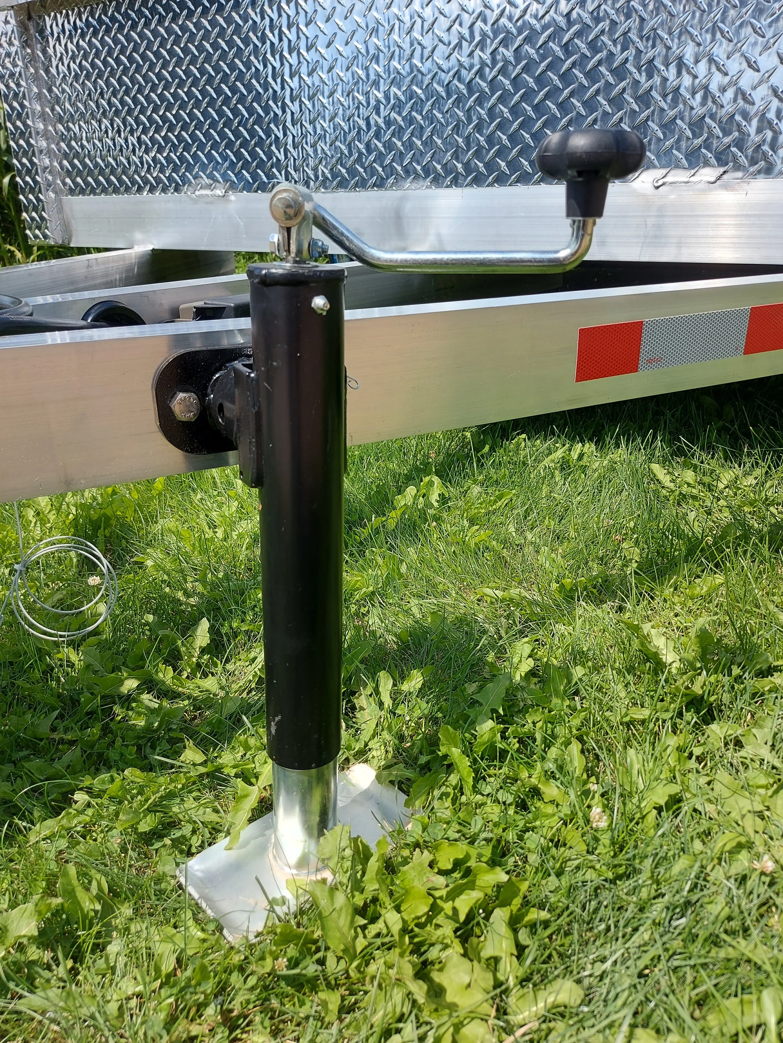 Close-up of a boat trailer jack stand on grass, with a black pole, a silver base, and a handle on the side of the trailer support arm, and a diamond plate section of the trailer in the background.