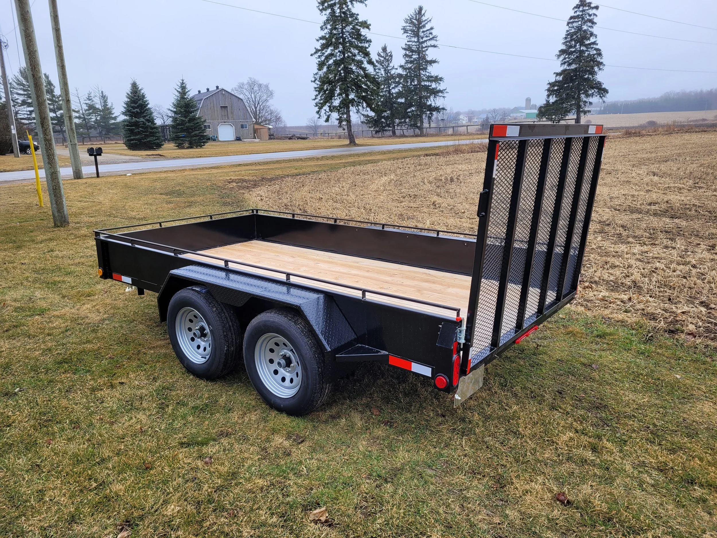 Black flatbed utility trailer with a fold-up ramp gate, parked on a grassy area near a road.