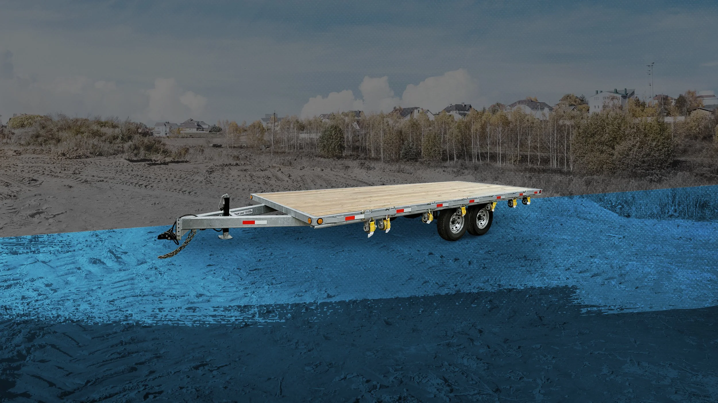 A flatbed trailer with two axles and a wooden deck parked on a blue painted surface outdoors, with a background of a dirt field, trees, and houses under a partly cloudy sky.