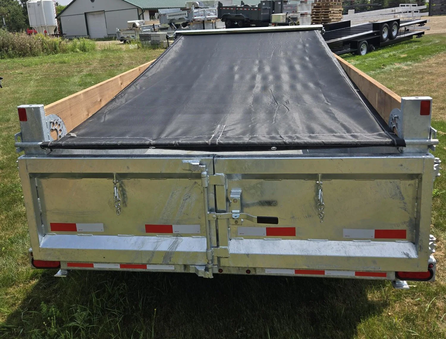 A flatbed trailer with a black tarp cover, parked on grass in an outdoor area with other trailers and buildings in the background.
