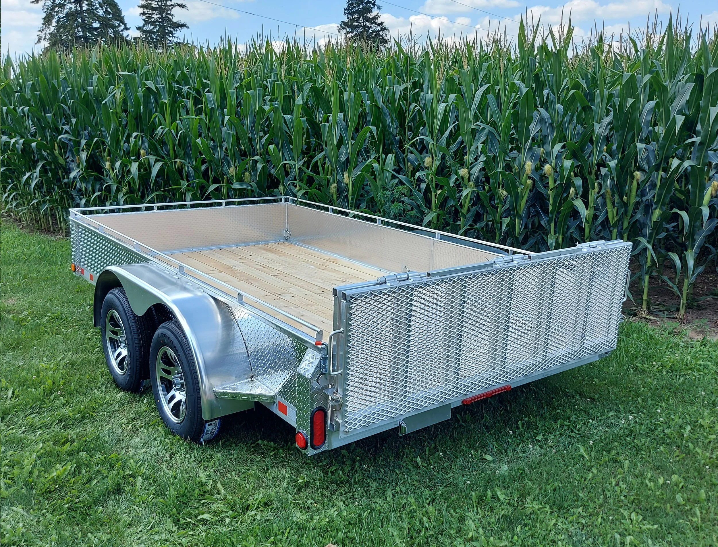 Empty metal utility trailer with wood floor parked on grass beside a cornfield.