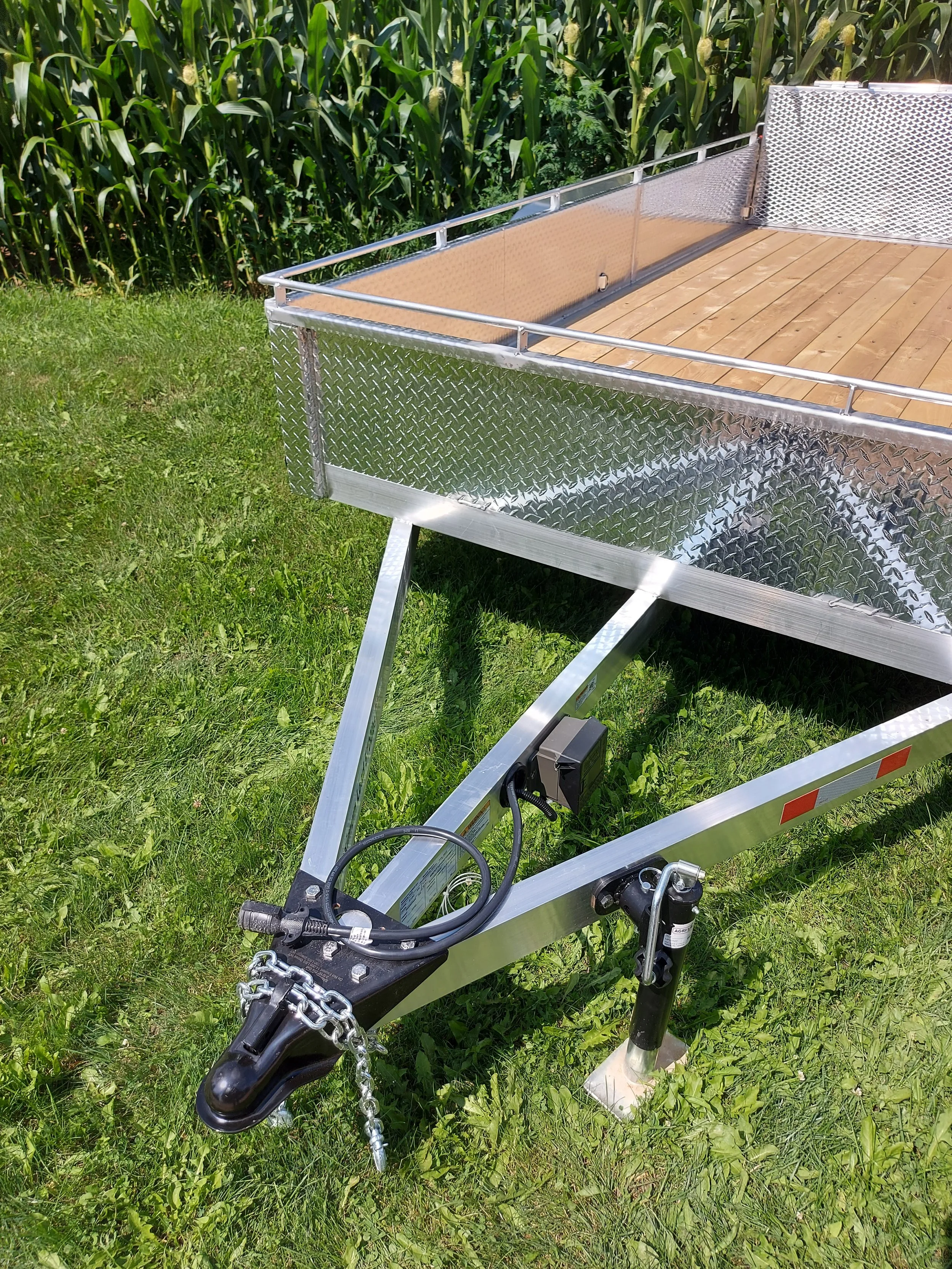 A utility trailer with a wooden floor, metallic sides, and diamond plate edges, parked on grass with a green cornfield in the background.