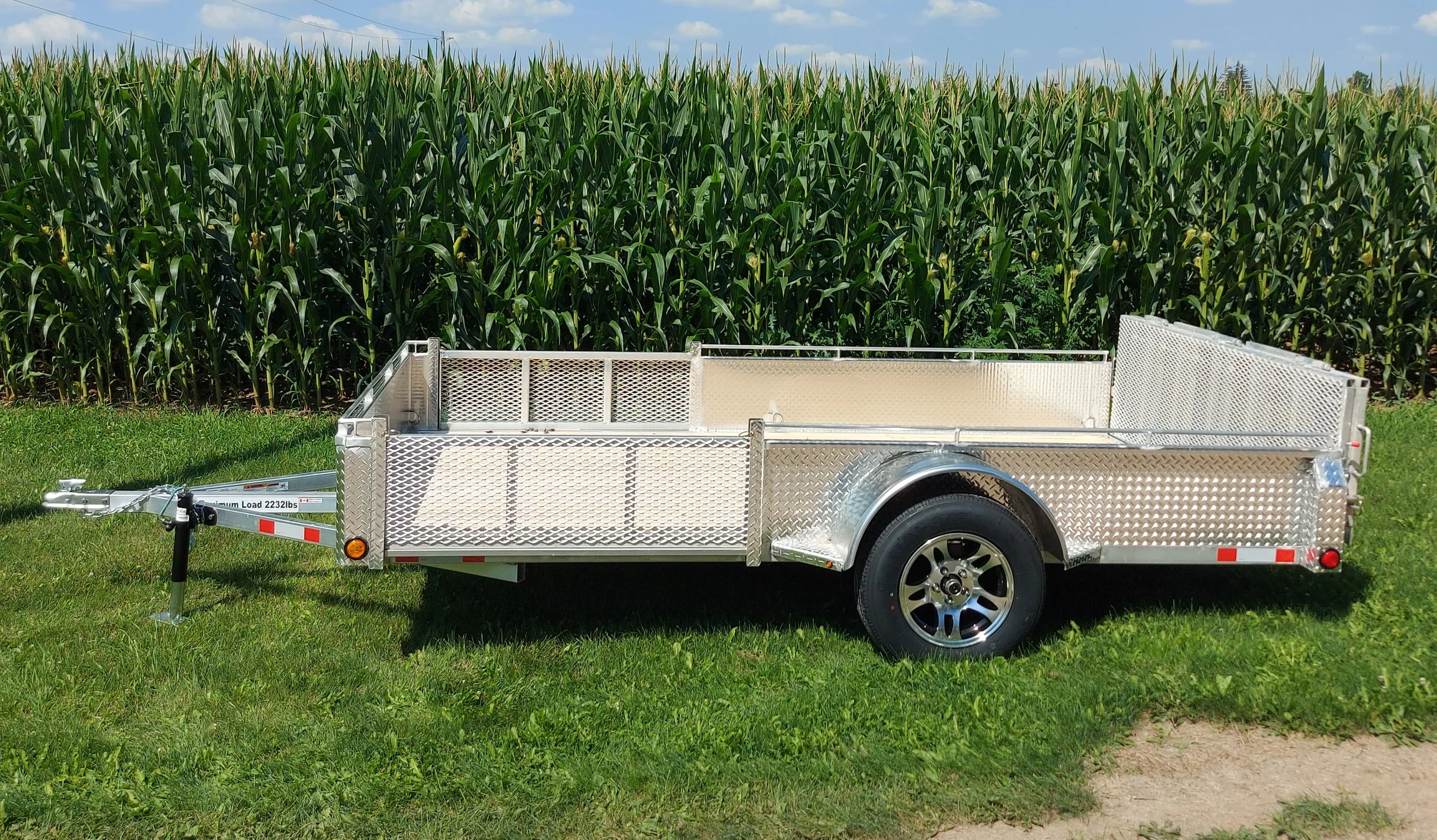An aluminum utility trailer parked on grass in front of a cornfield
