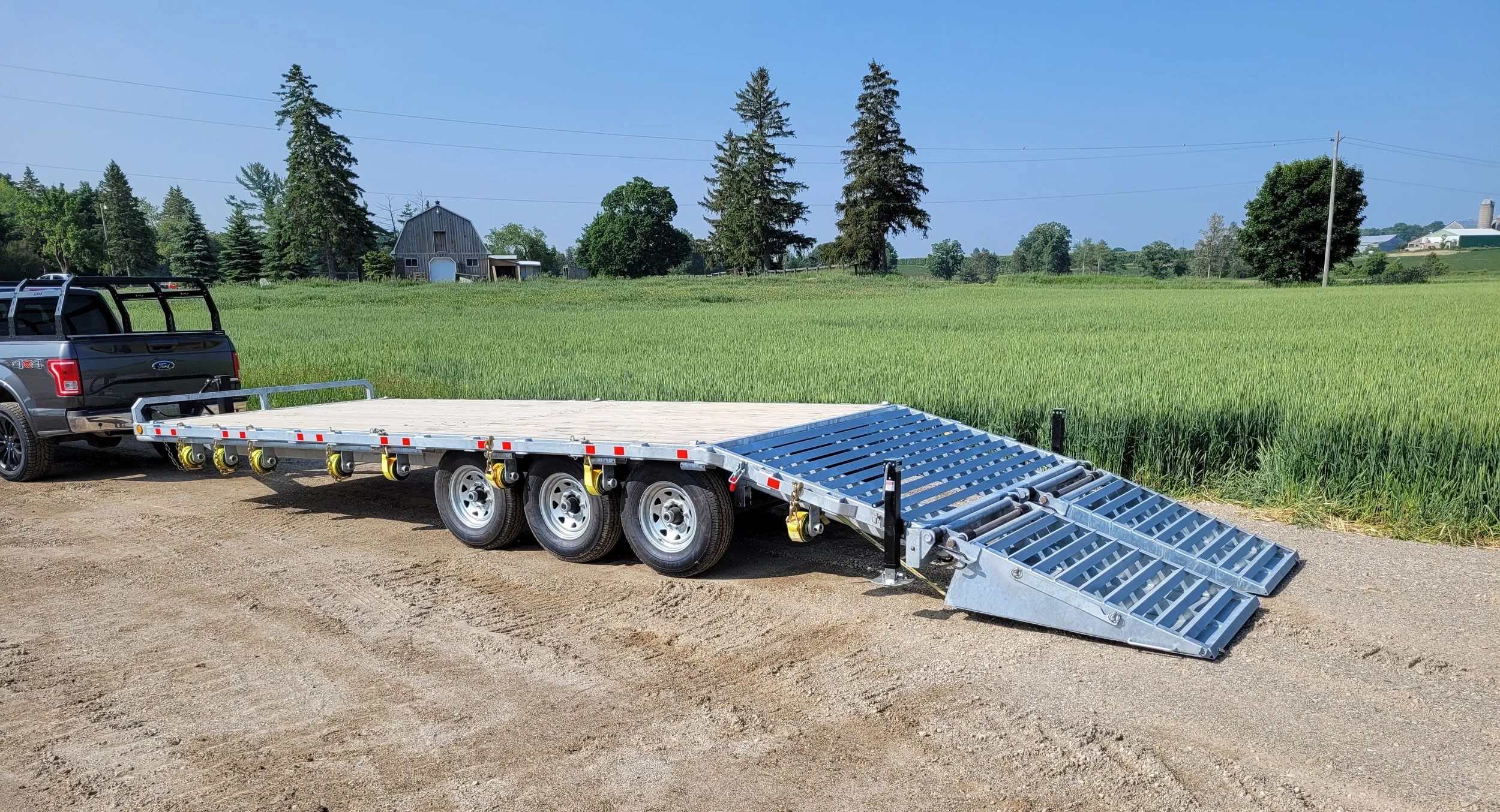 A black pickup truck hooked to a flatbed trailer with a metal loading ramp, parked on a dirt road in front of a green field and farm buildings.