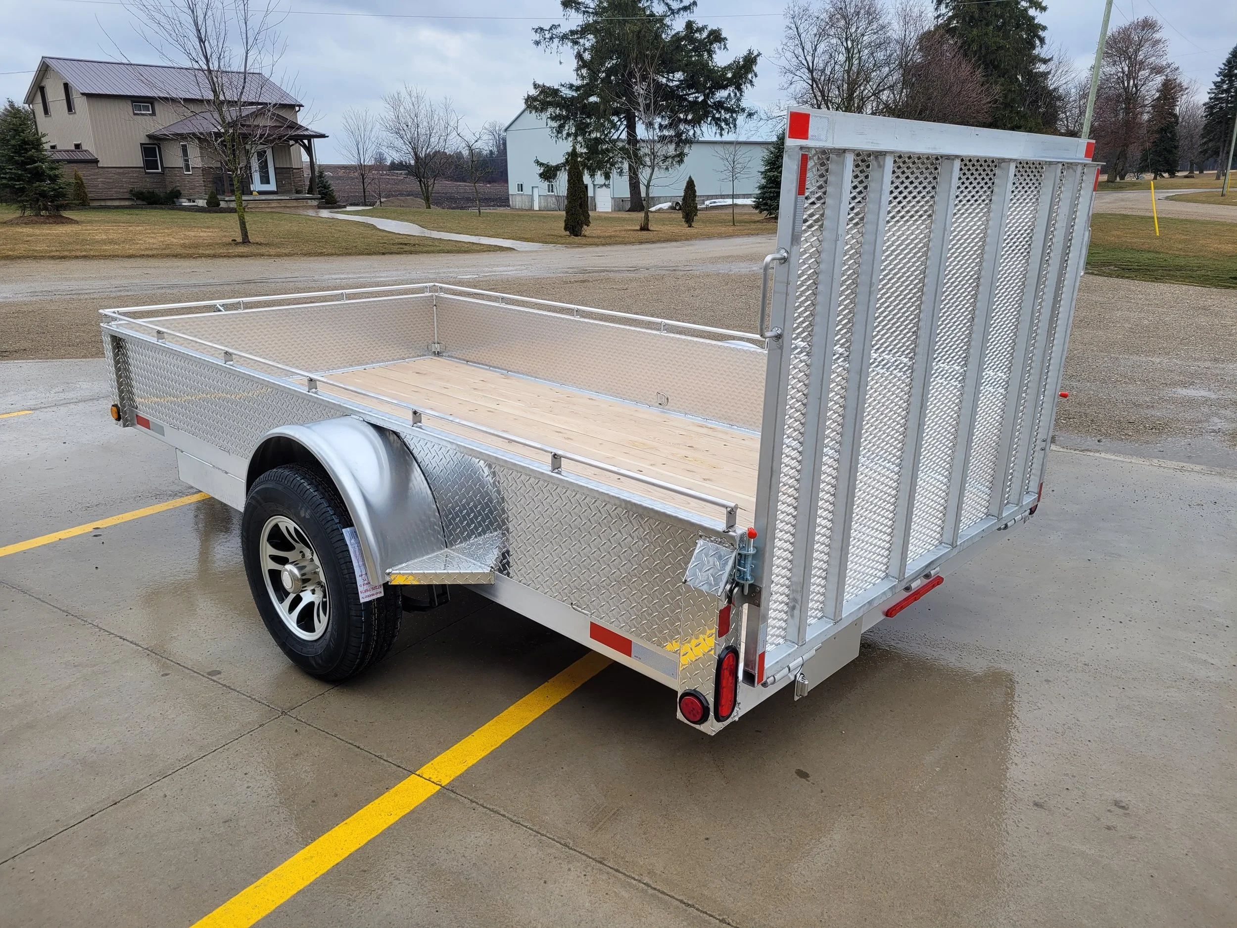 A silver utility trailer with a wooden floor, parked in a lot, with an open ramp at the back for unloading.