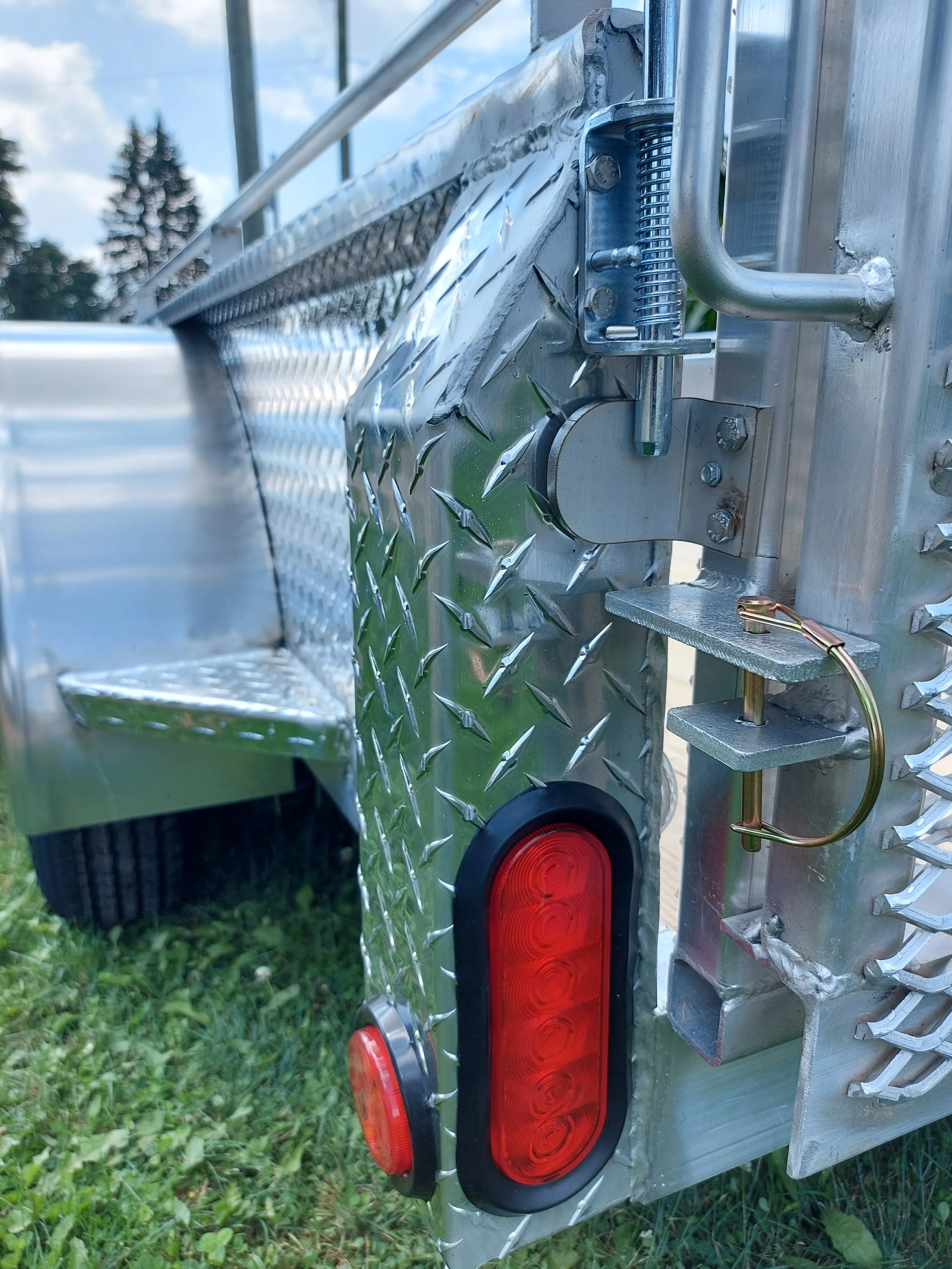 Close-up of the rear corner of a shiny, diamond-plate metal fire truck, showing a red oval taillight and a small red reflector on the grass.