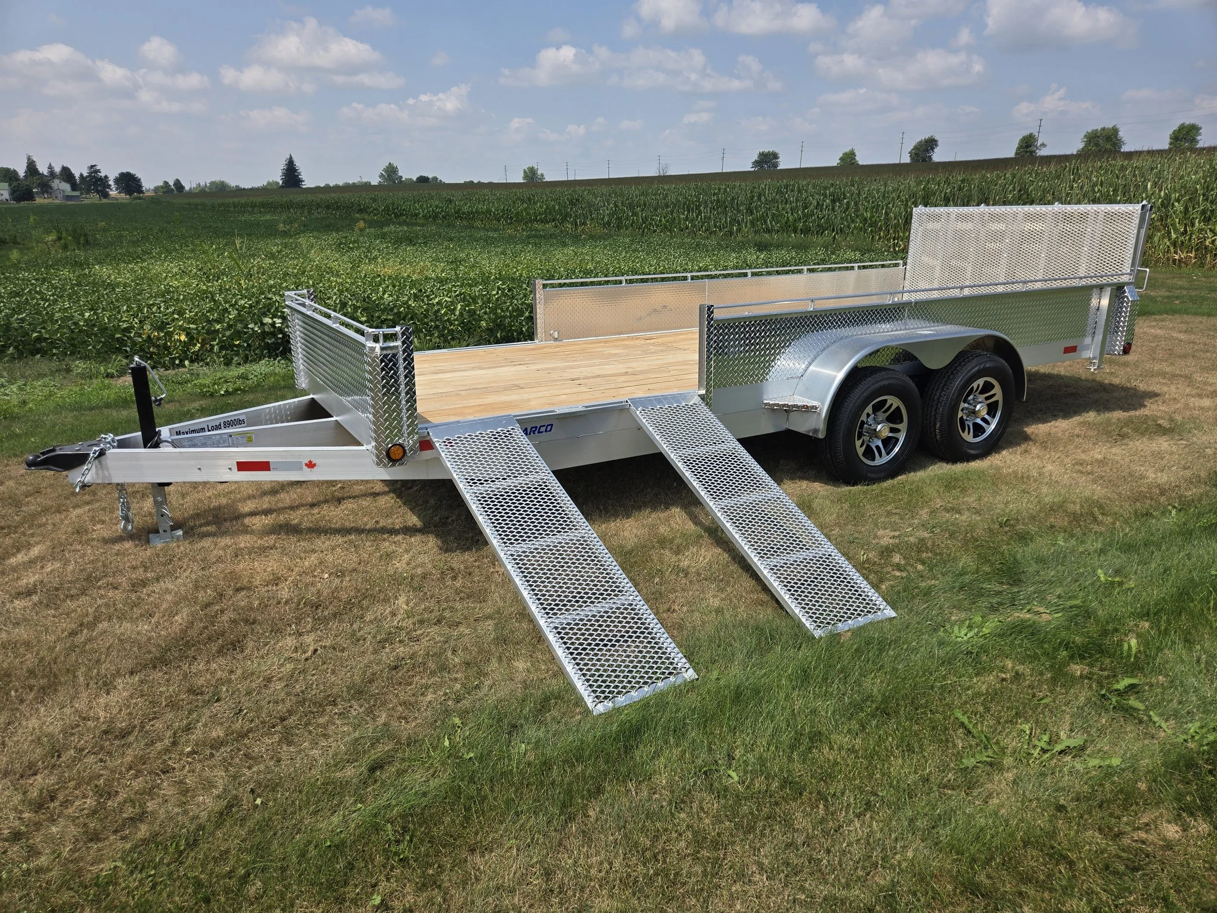 A flatbed utility trailer with a metal frame, two axles with black tires, and a loading ramp in a grassy field near a farm with crops under a partly cloudy sky.