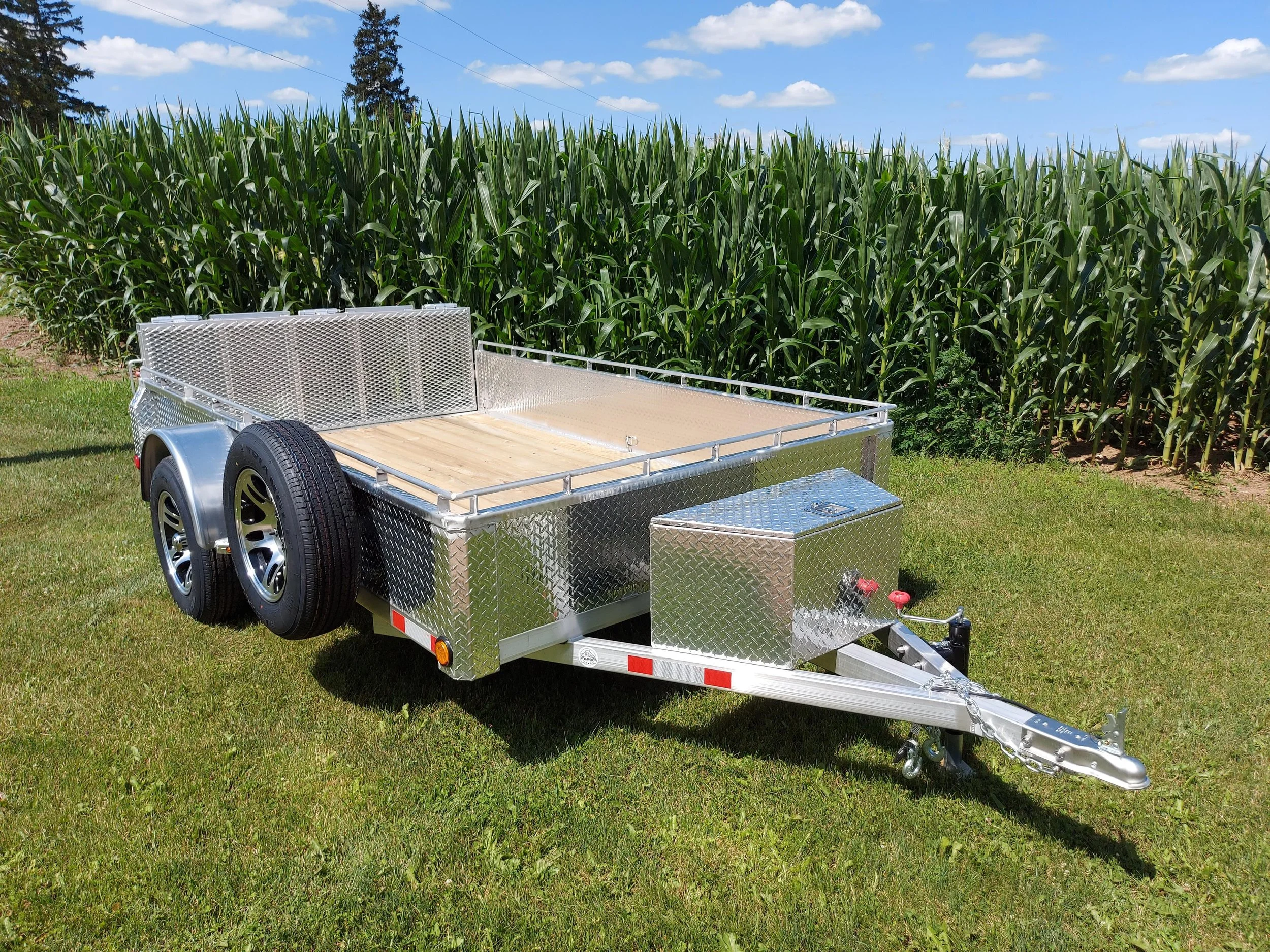 Silver flatbed utility trailer with wooden floor parked on grass in front of a cornstalk field under a blue sky with clouds.