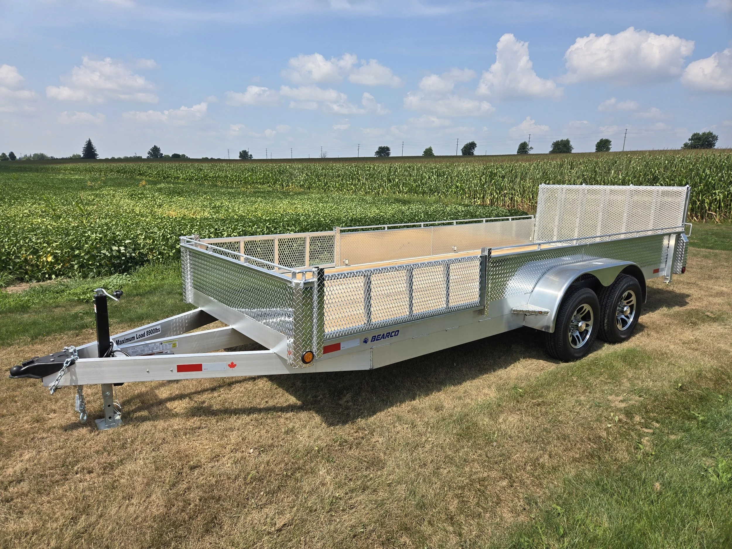 Empty flatbed utility trailer parked on grass field, with green crops and blue sky with clouds in background.