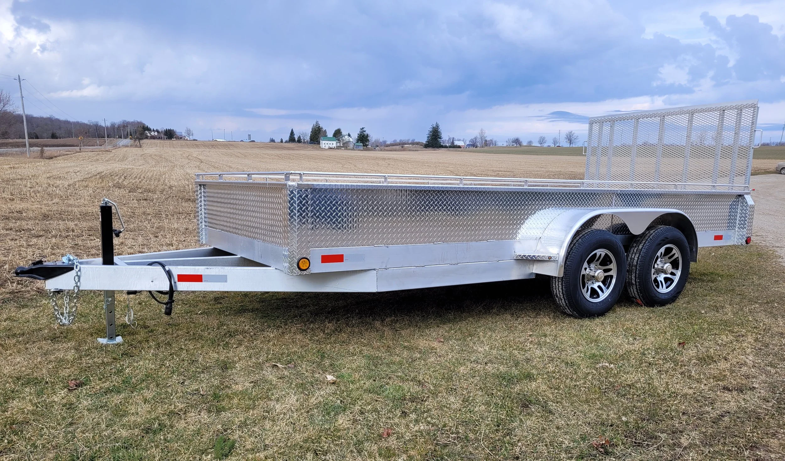 A silver utility trailer parked on grass in an open field, with a partly cloudy sky above.