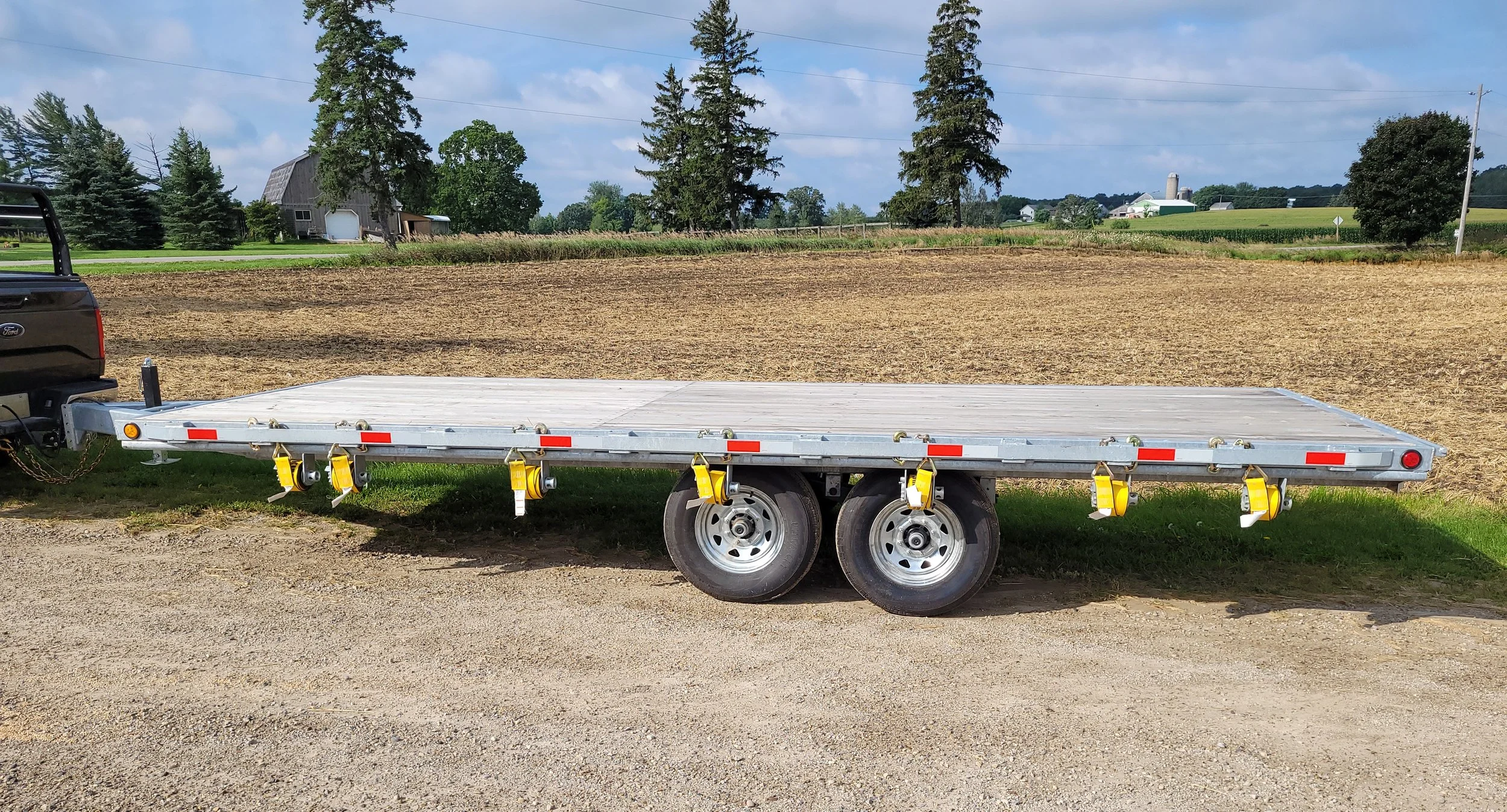 Empty flatbed trailer attached to a pickup truck, parked on a gravel area near a farm field with a barn, trees, and farm buildings in the background.