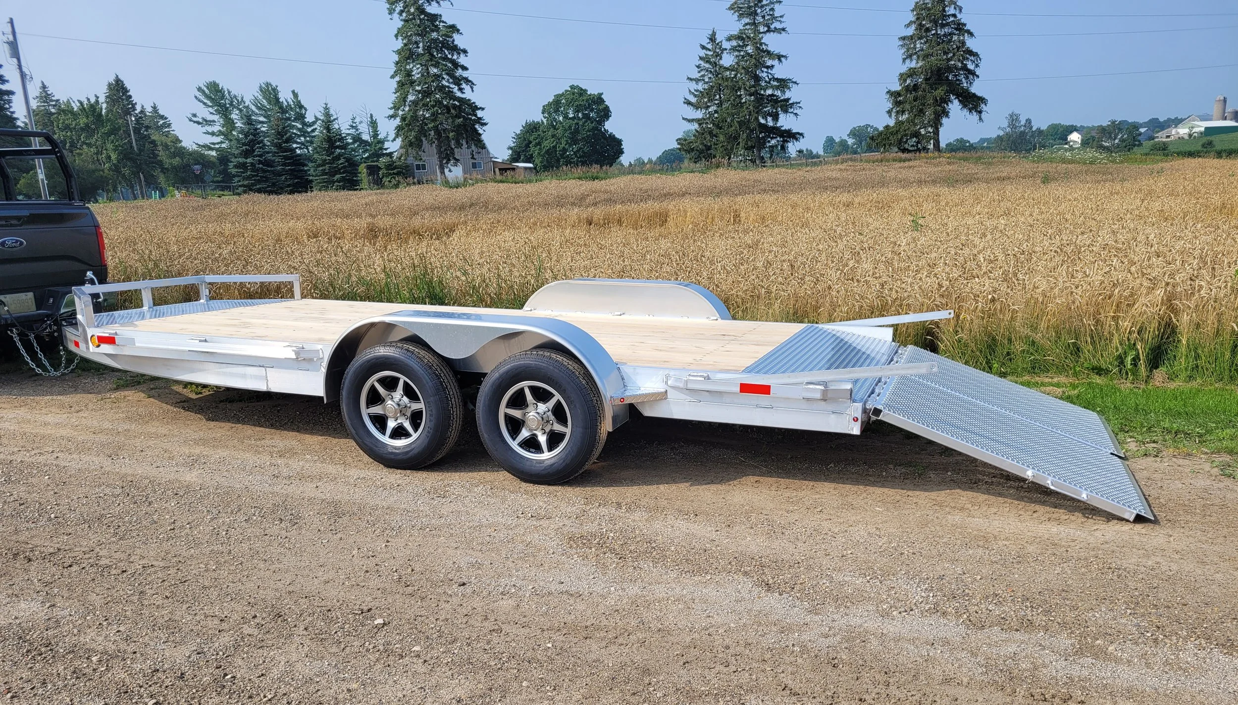 Silver dual-axle car trailer attached to a black pickup truck, on a dirt ground near a field of tall grass or grain, with trees and houses in the background.