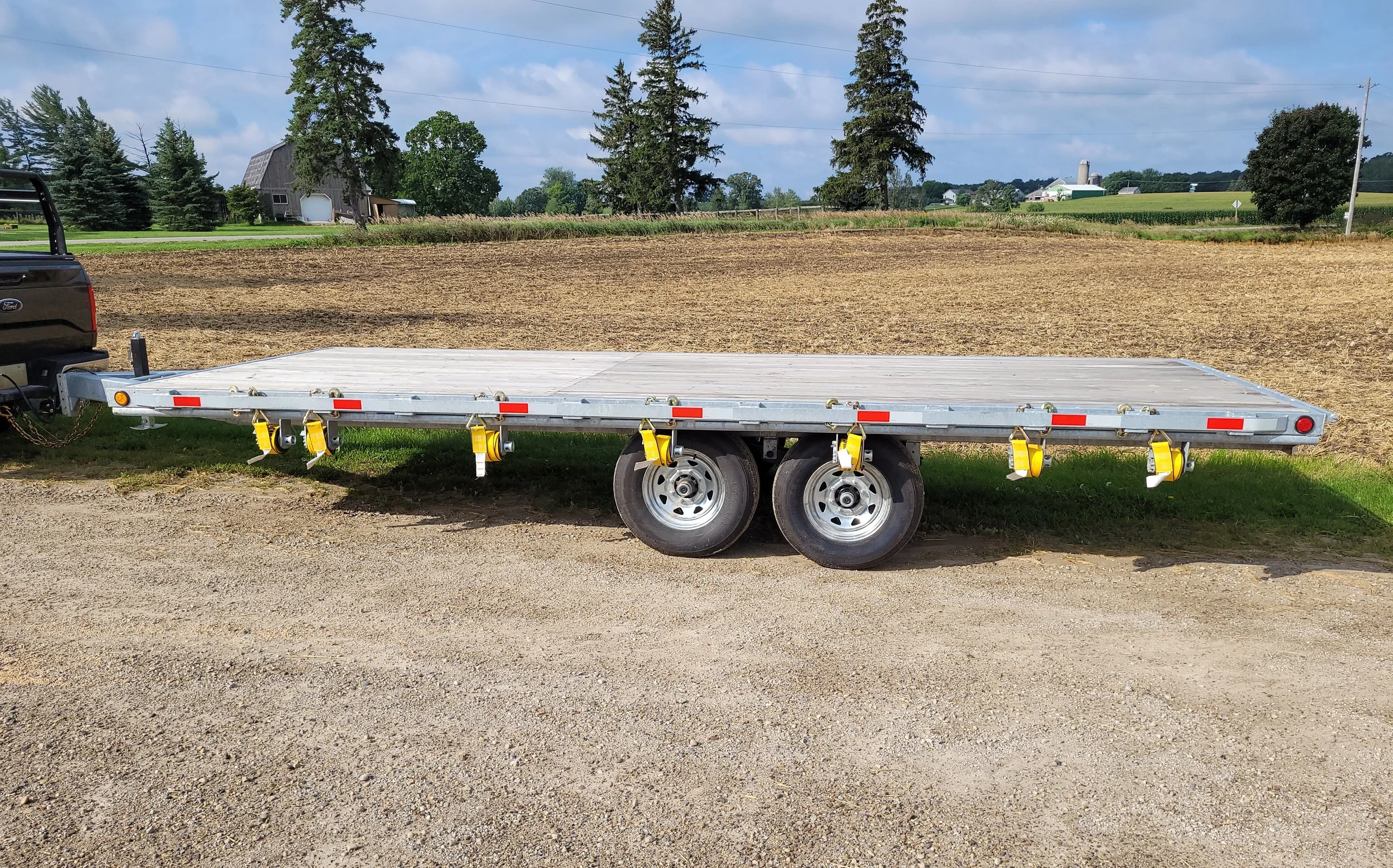 A flatbed trailer parked on a gravel lot, attached to a vehicle outside in a rural area with fields, trees, barns, and farm buildings in the background.