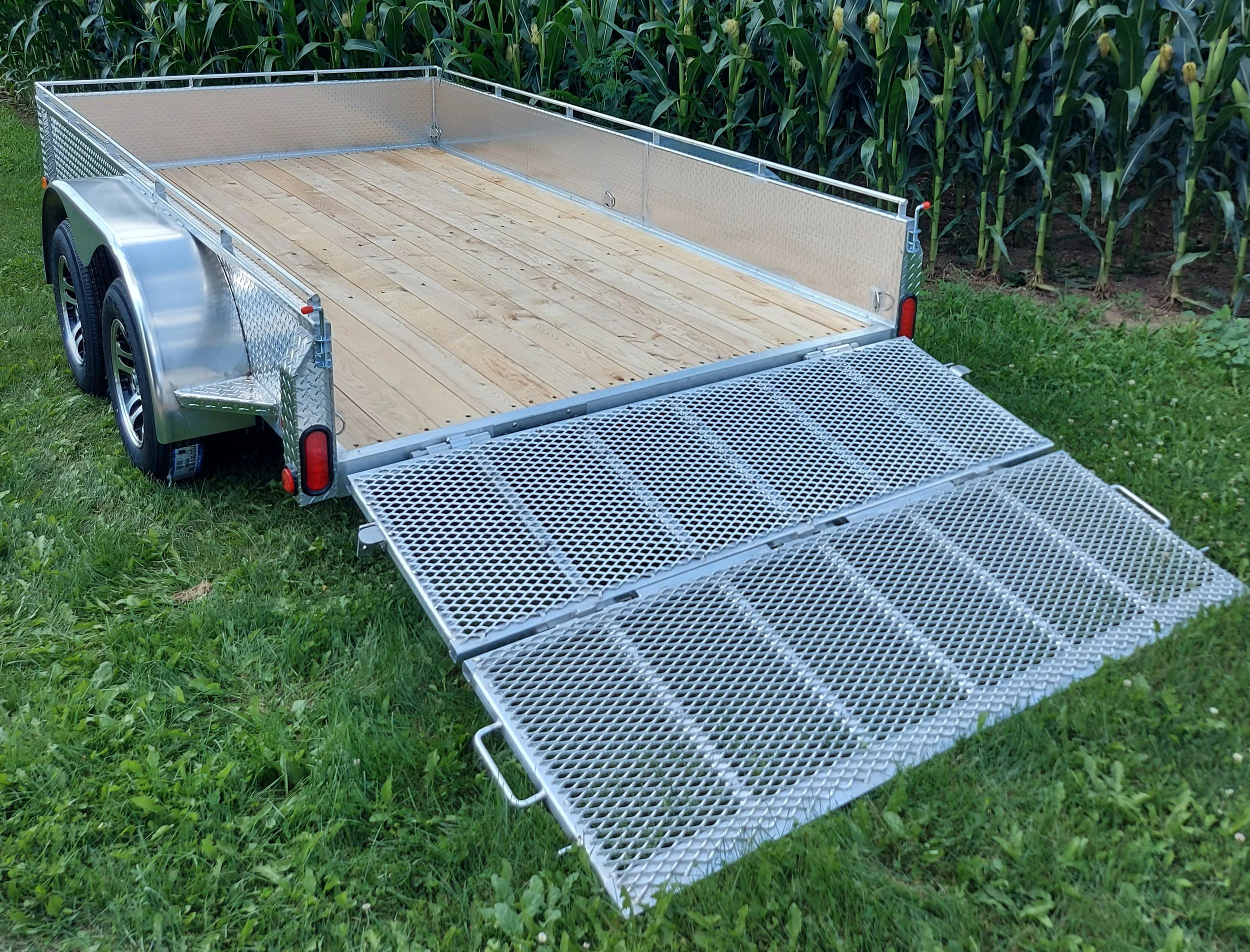 A flatbed trailer with a metal frame and wooden flooring parked on grass in front of a cornfield.