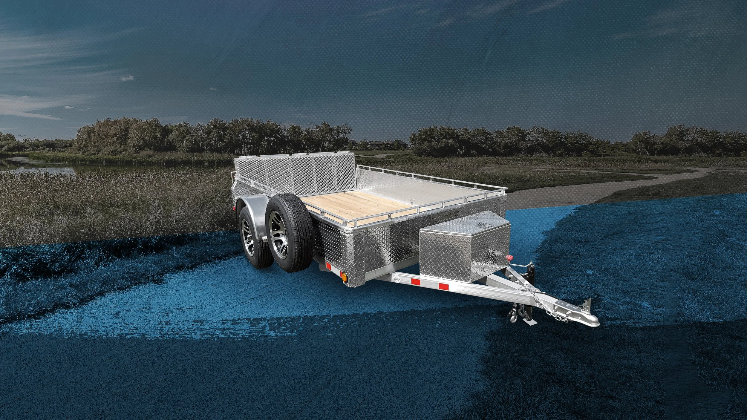 A silver utility trailer with two tires parked on a grassy field near a body of water under a partly cloudy sky.