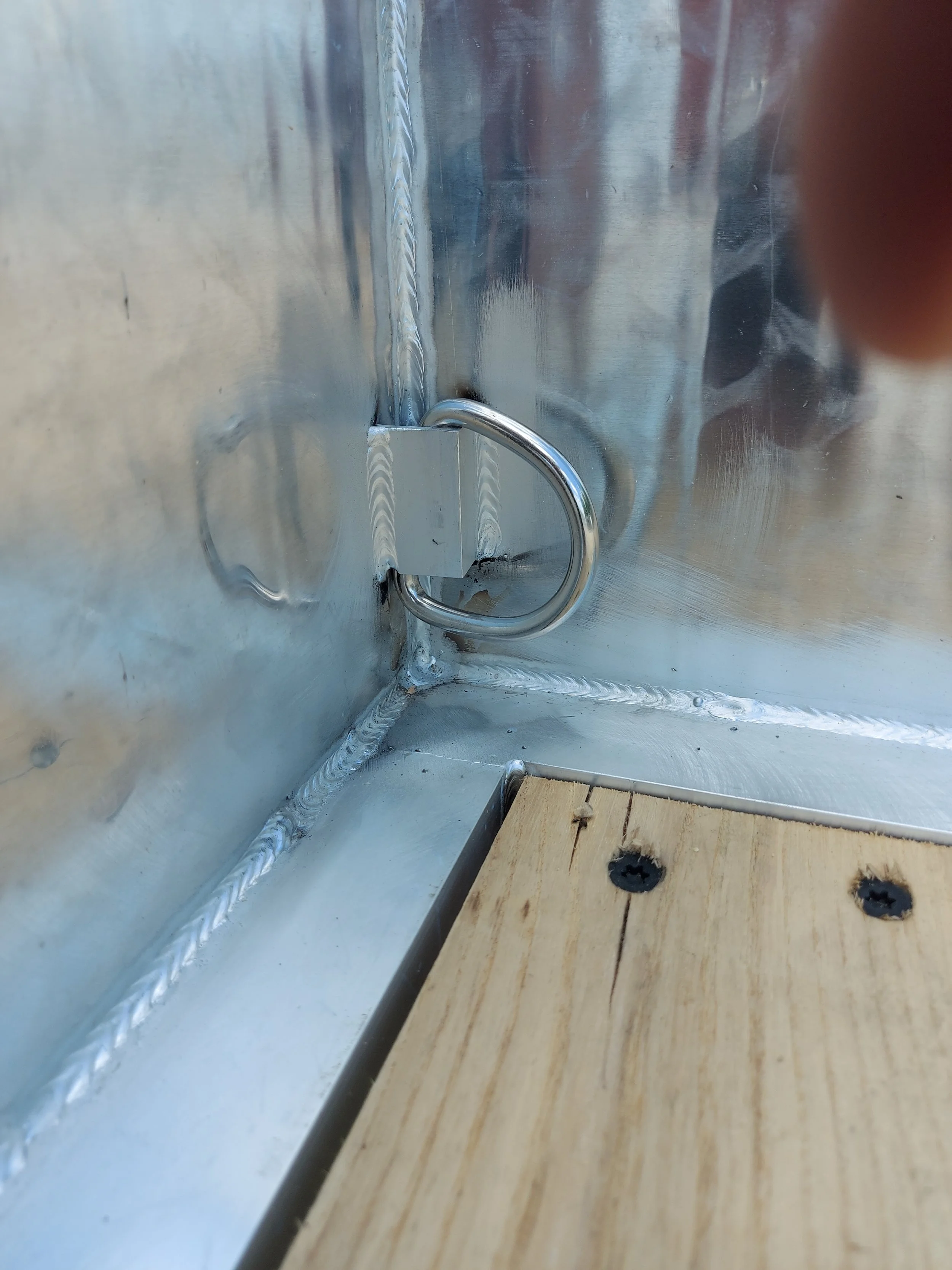Close-up of a metal latch attached to a corner of a metal structure with a wooden surface next to it, with visible welds and screws.