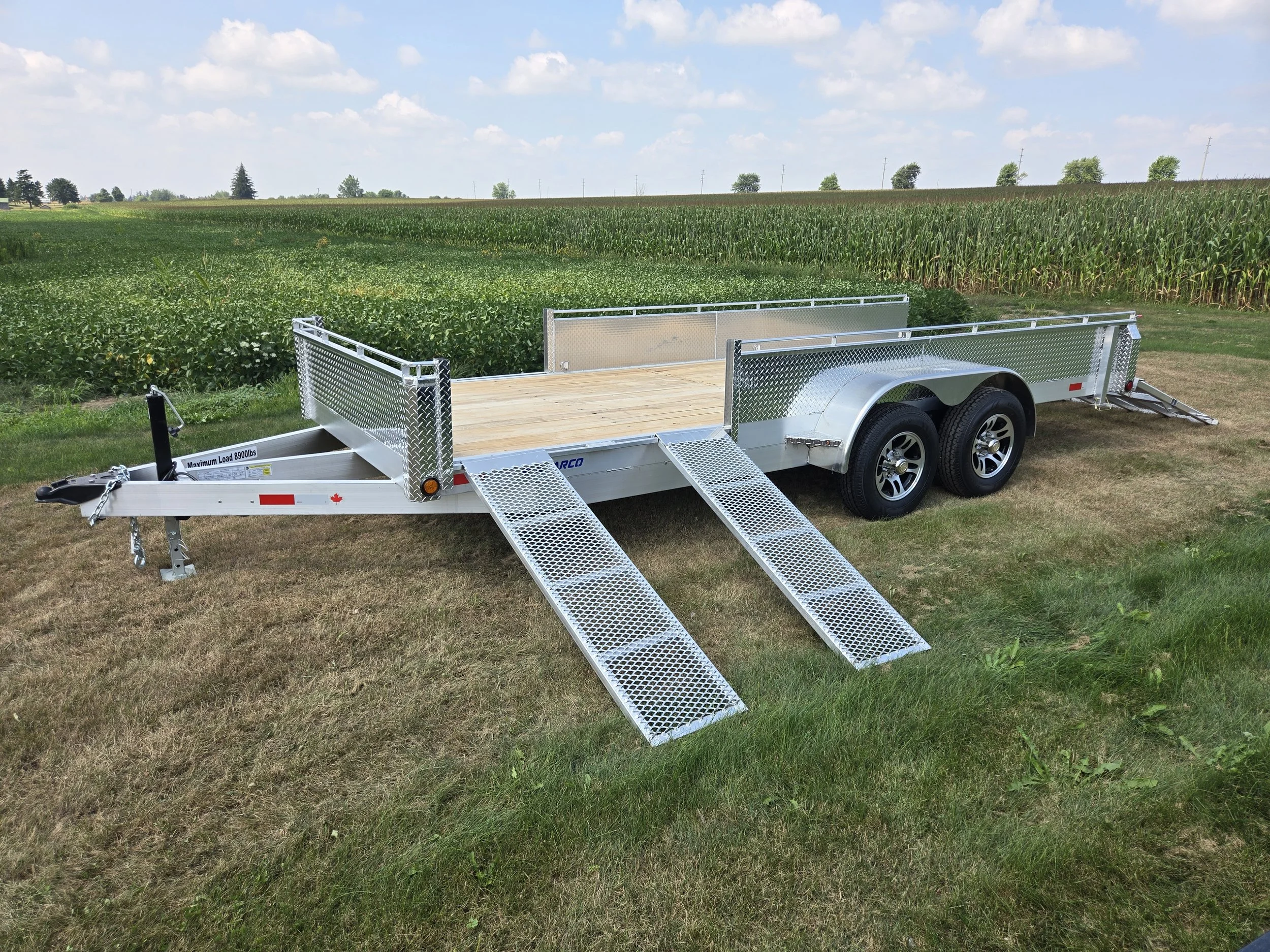 A flatbed utility trailer with dual axles and metal ramps, parked on grass with a field of crops and a partly cloudy sky in the background.