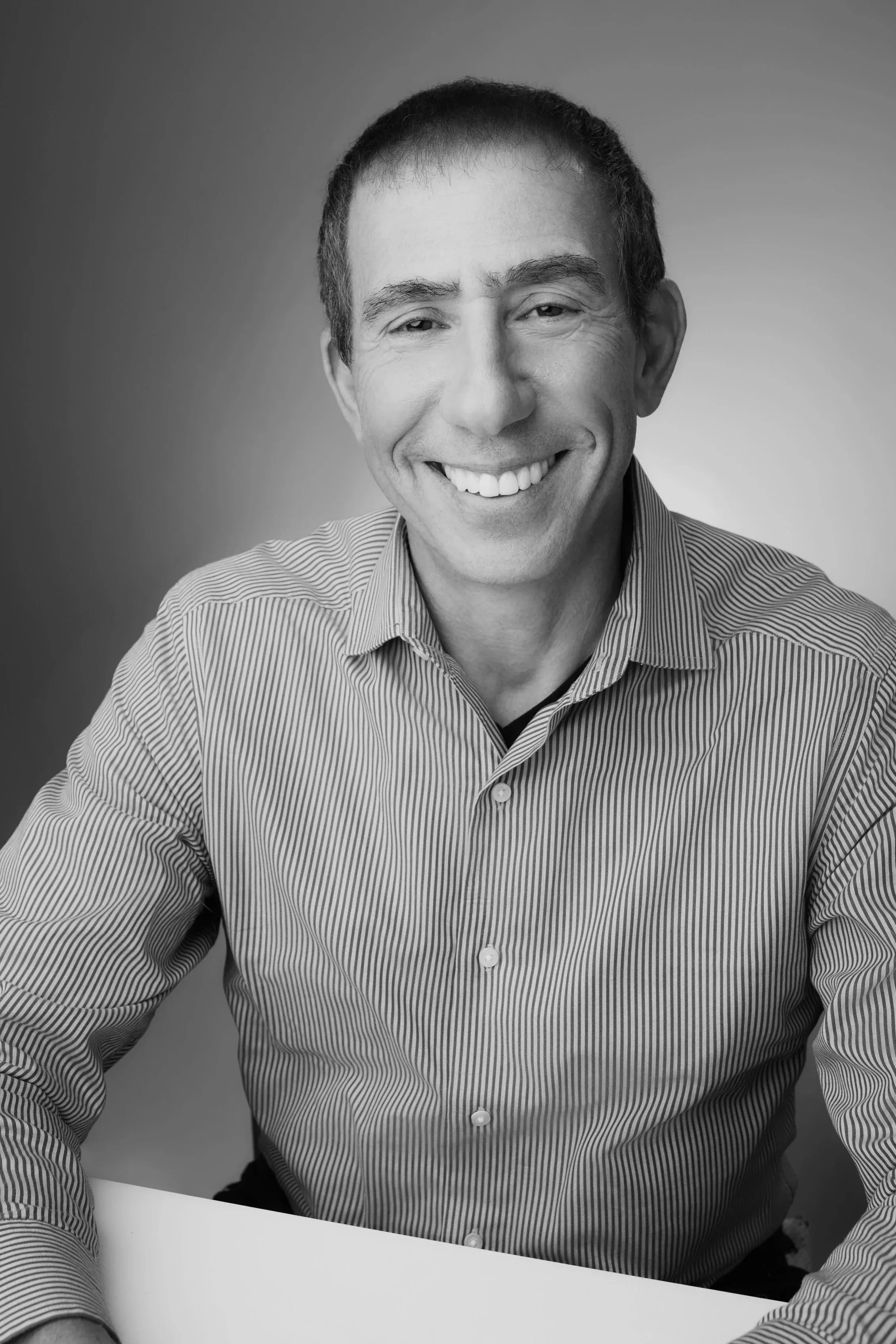 A smiling man with short dark hair wearing a striped collared shirt, sitting at a table, in a black-and-white portrait.