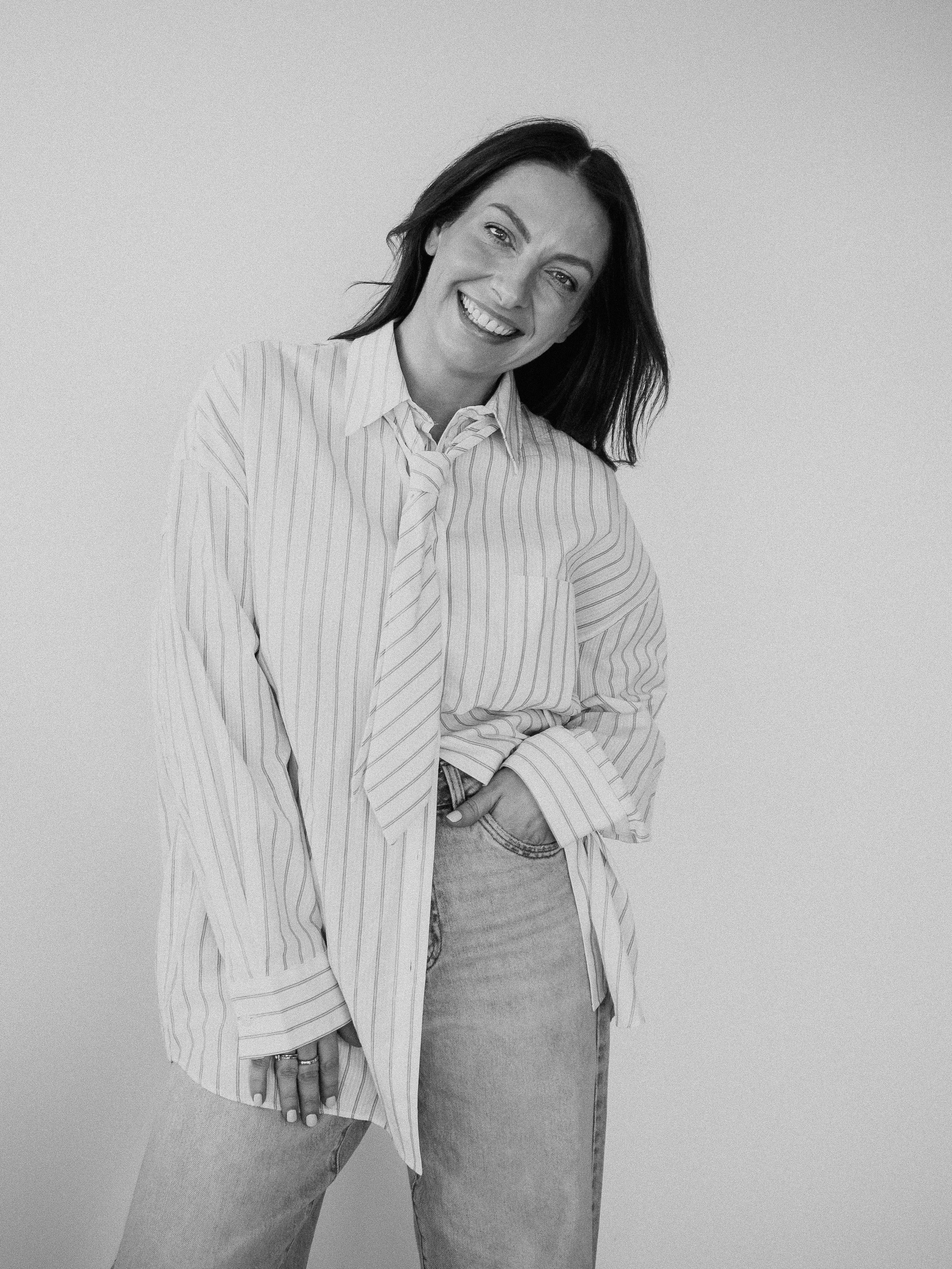 A woman with shoulder-length dark hair smiling and wearing a striped shirt with a tie, jeans, and rings on her fingers, standing against a plain light-colored background.
