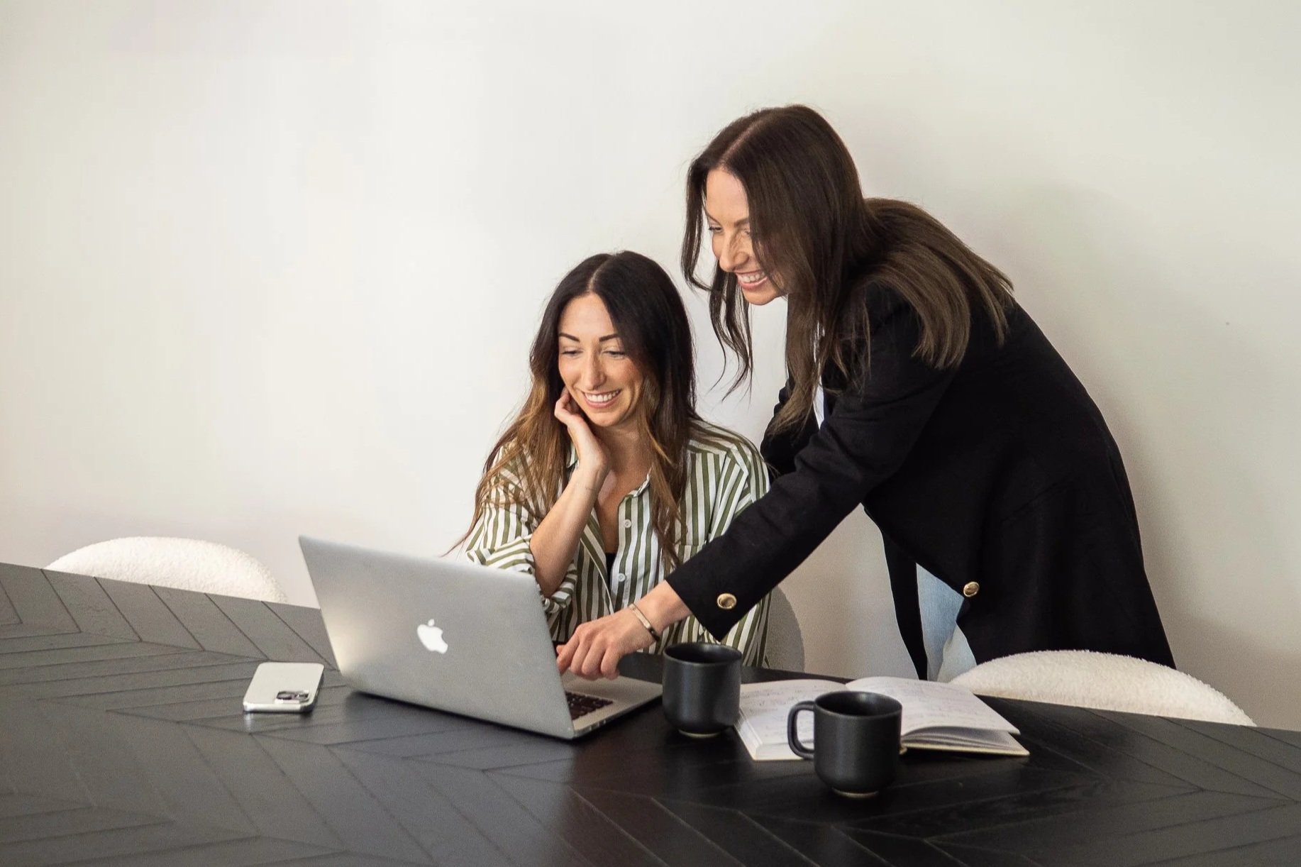 Two women working together at a black table with laptops and coffee mugs, smiling and looking at the screen.