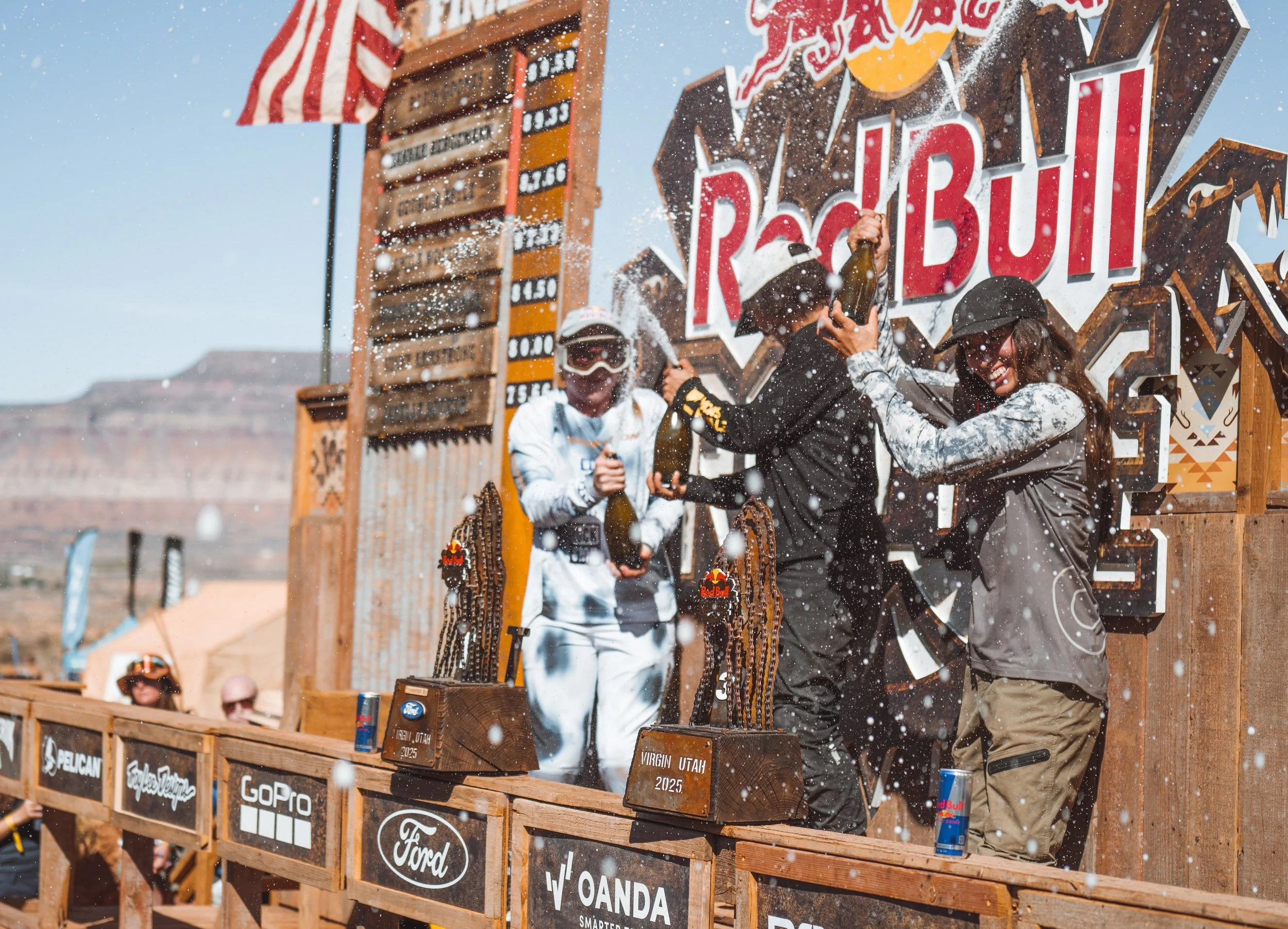 People celebrating on a podium at a Red Bull event with champagne, surrounded by sponsorship banners, in an outdoor desert location.