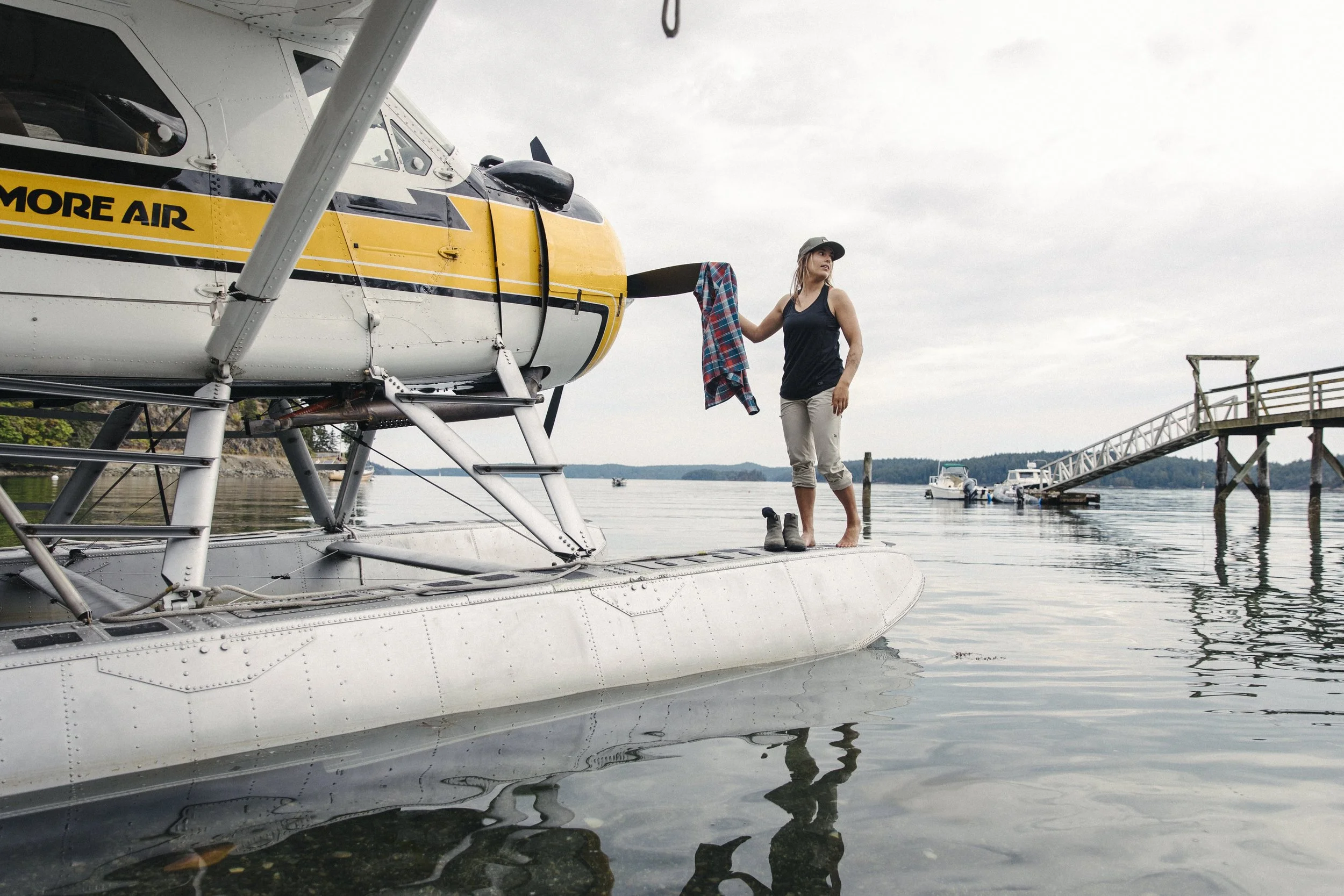 A woman standing on the float of a seaplane near a dock, holding a plaid shirt, with boats and a wooden pier in the background.