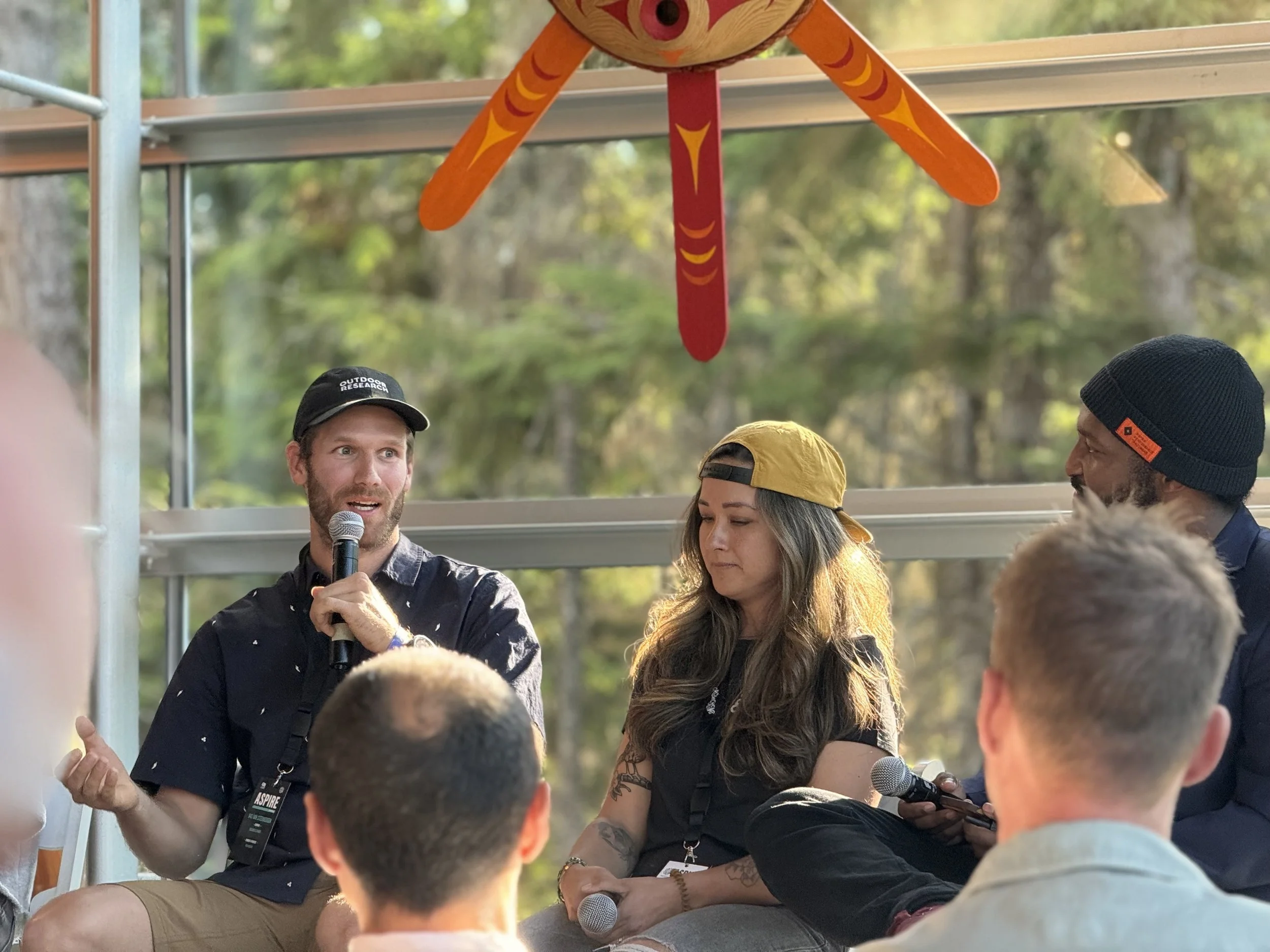 Group of people sitting on a stage outdoors during a panel discussion, with a large dragon-shaped puppet hanging above them, surrounded by trees.