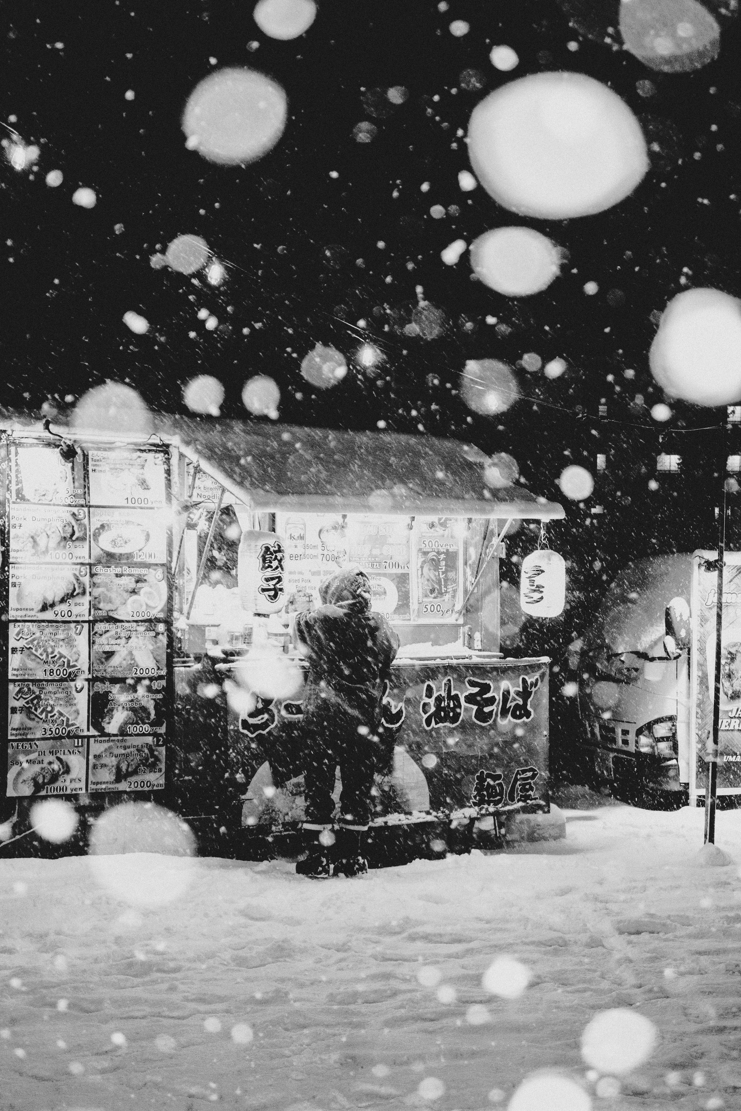 A person at a food stand in a snowy outdoor scene, with falling snowflakes and illuminated signs in the background.