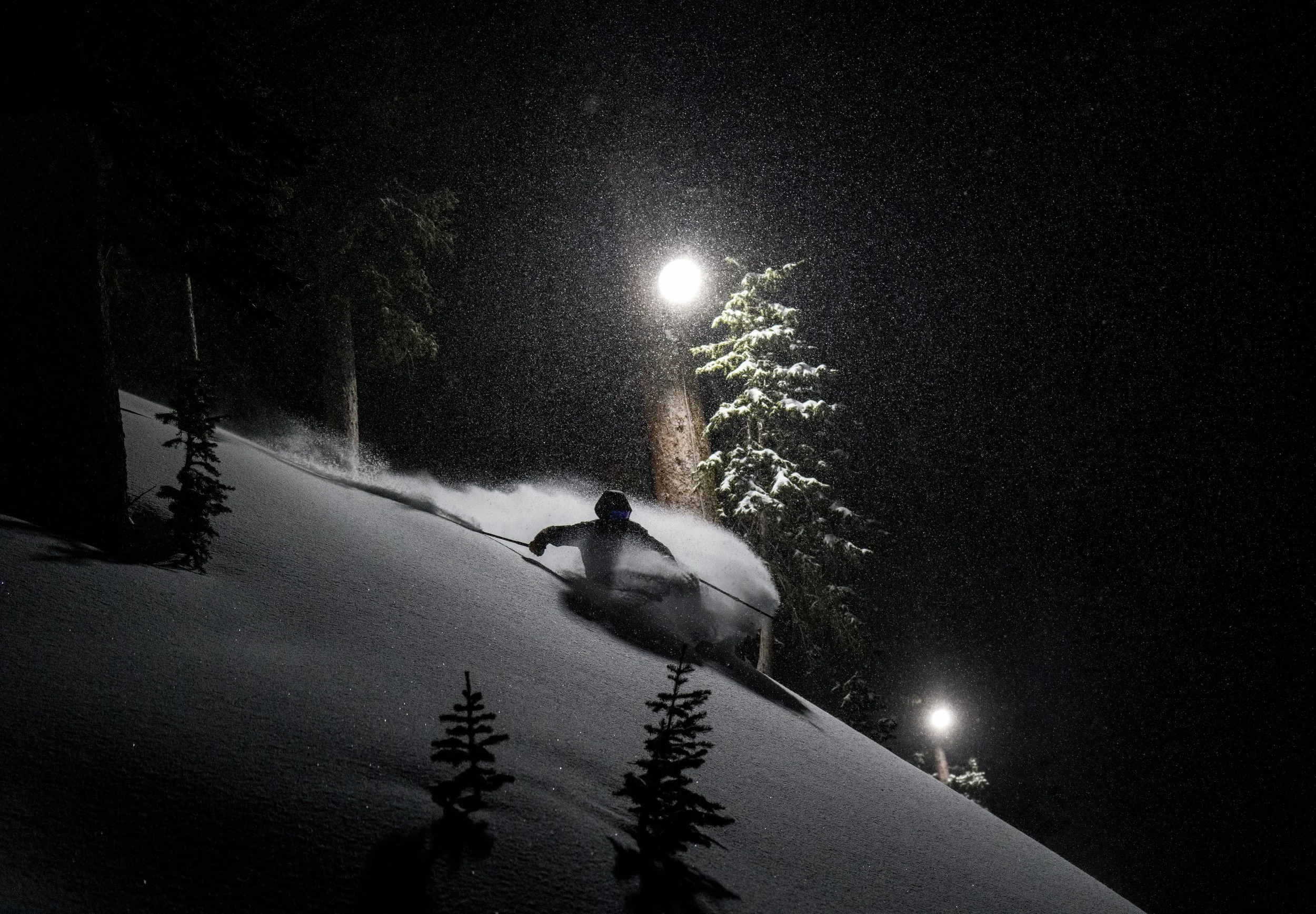 A skier wearing nighttime gear is skiing down a snow-covered slope at night under the moonlight, with snow falling around and trees in the background.