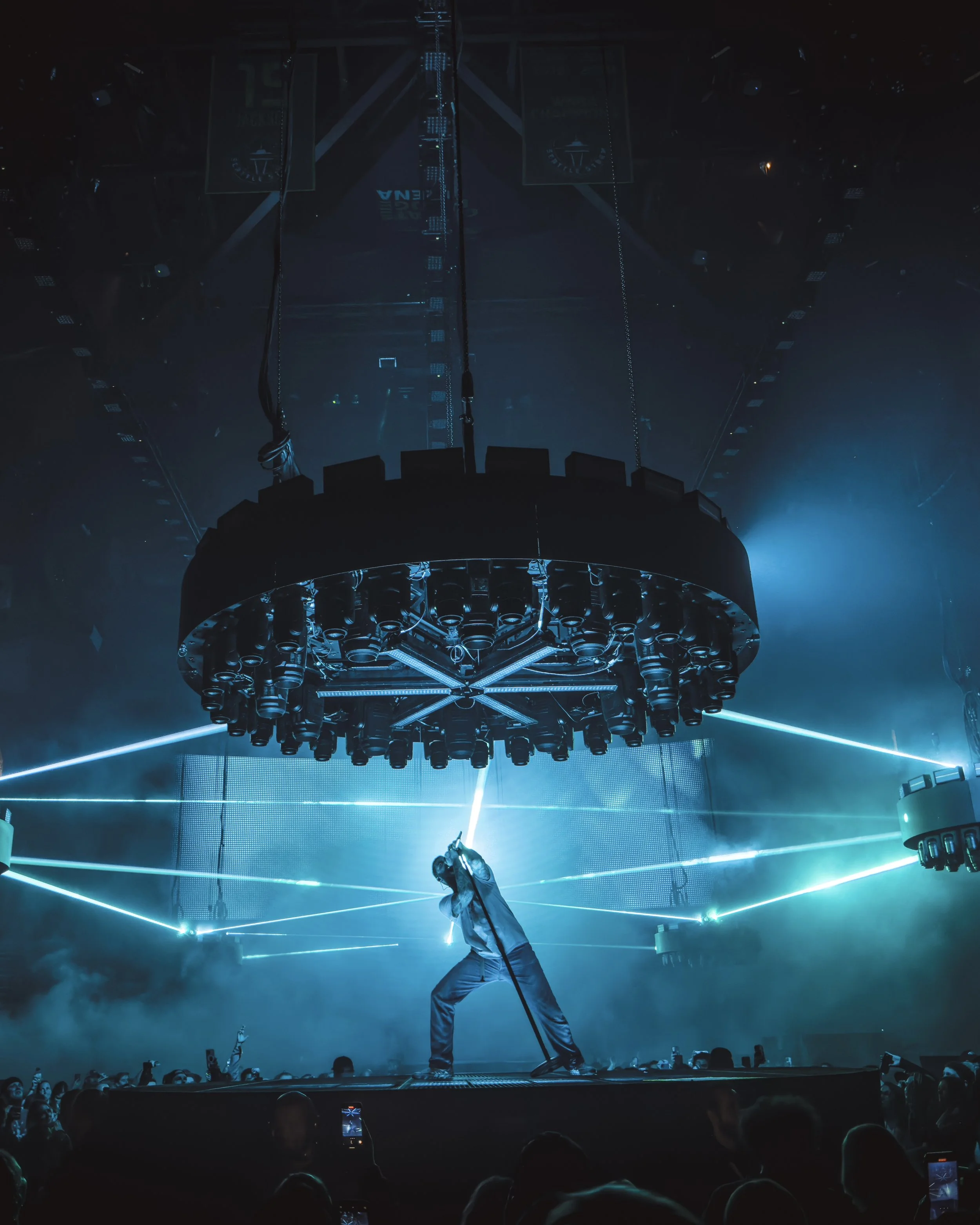 A performer on stage holding a microphone in a live concert with blue laser lights and a large circular lighting rig overhead, dark surroundings, and an audience in the foreground.