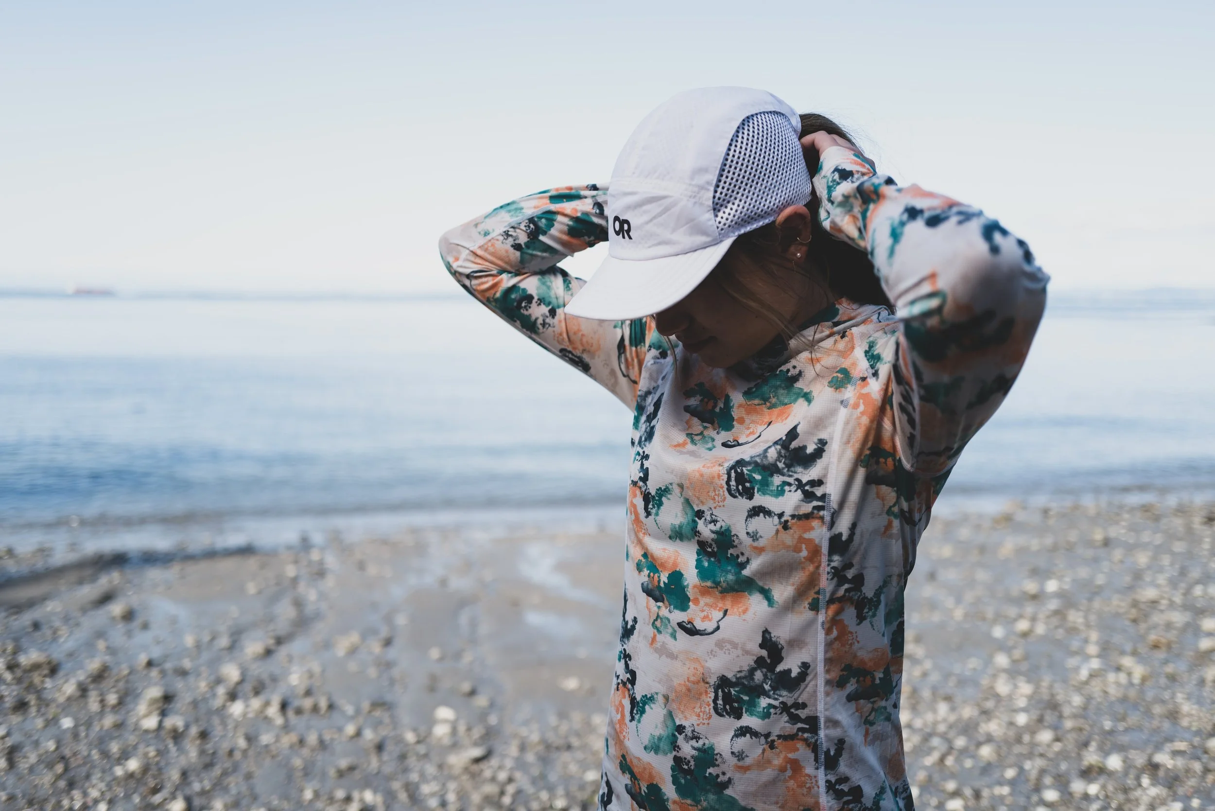 Woman in a floral patterned jacket and white cap adjusting her hat on a pebble-covered beach with calm water in the background.