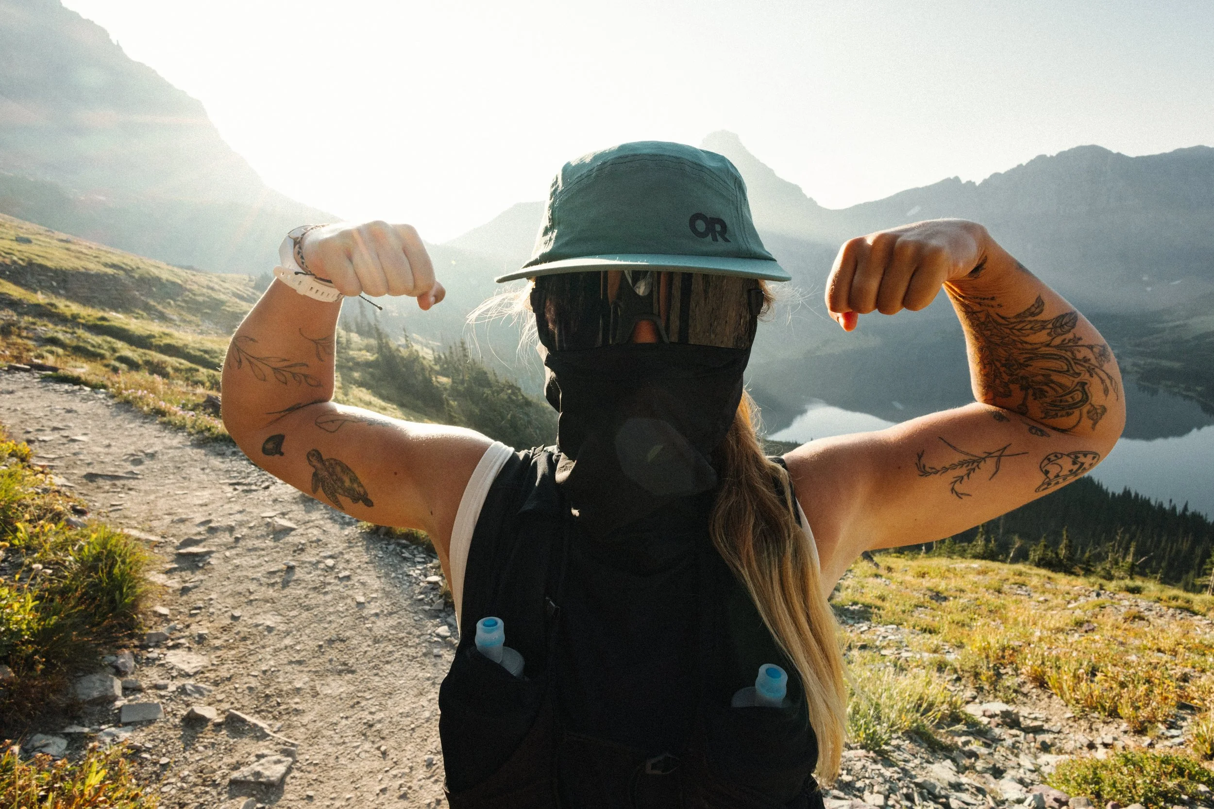 A woman with tattoos wearing a hat, sunglasses, and a mask, flexing her muscles on a mountainous trail near a lake during sunset.