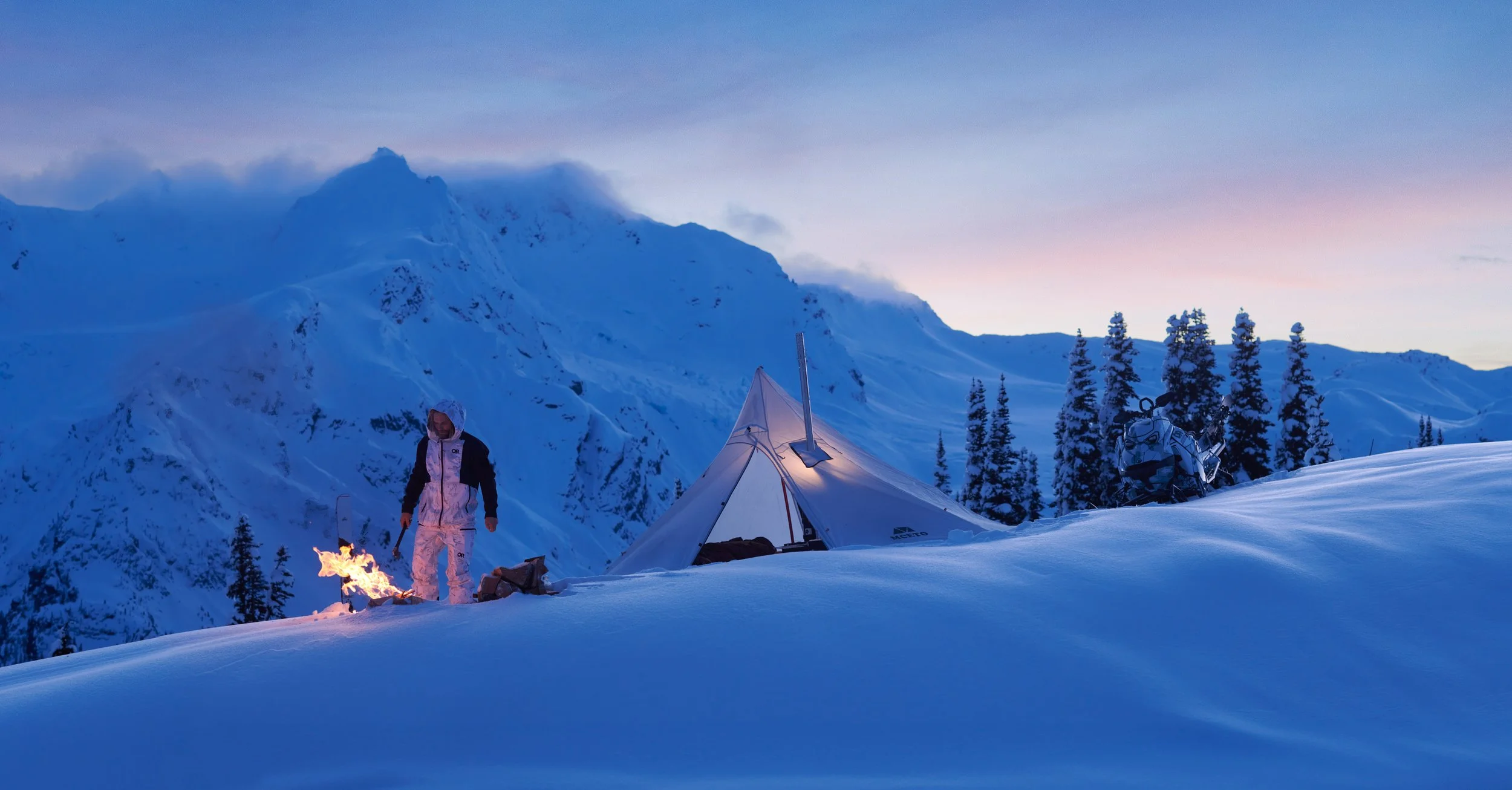 A person in winter gear near a campfire on snow with a tent and snow-covered mountains and trees in the background at dusk.