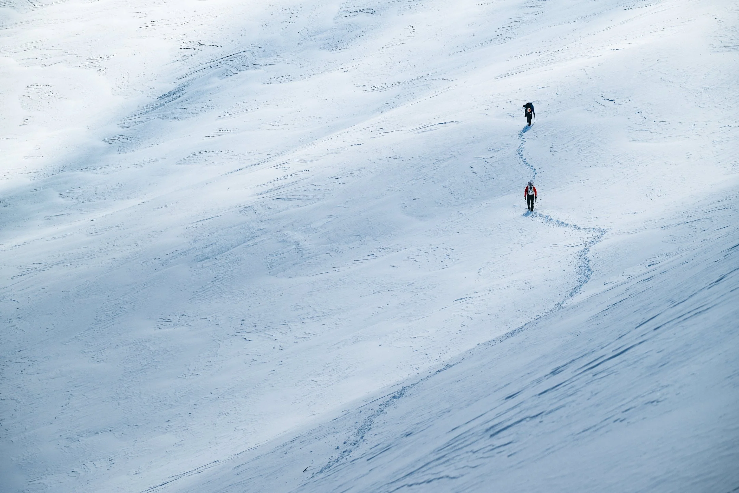 Two hikers trekking through snow on a mountain slope, creating footprints behind them.