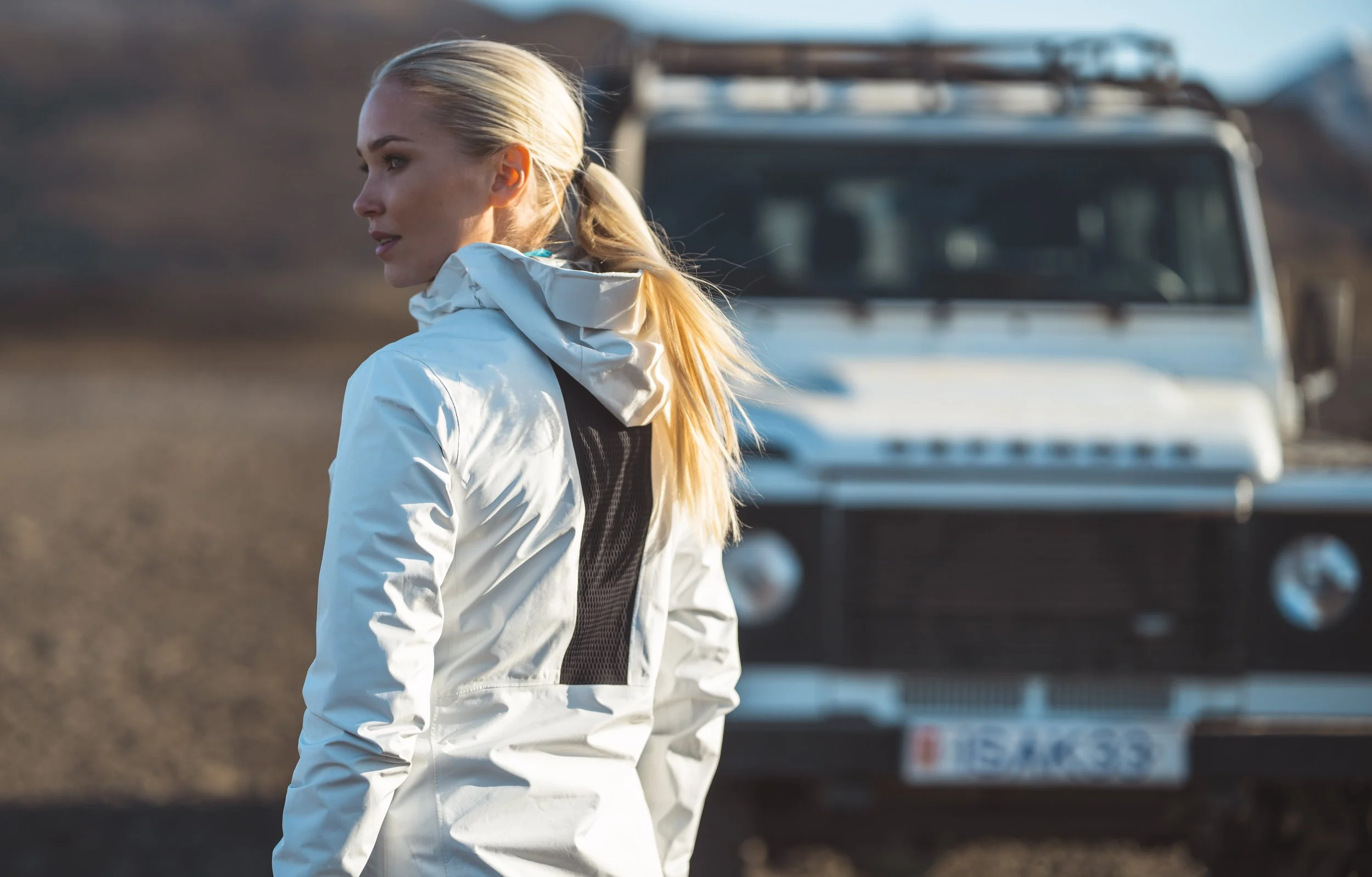 A woman with blonde hair tied in a ponytail, wearing a white jacket with a black mesh panel, stands outdoors with a vintage Land Rover in the background.