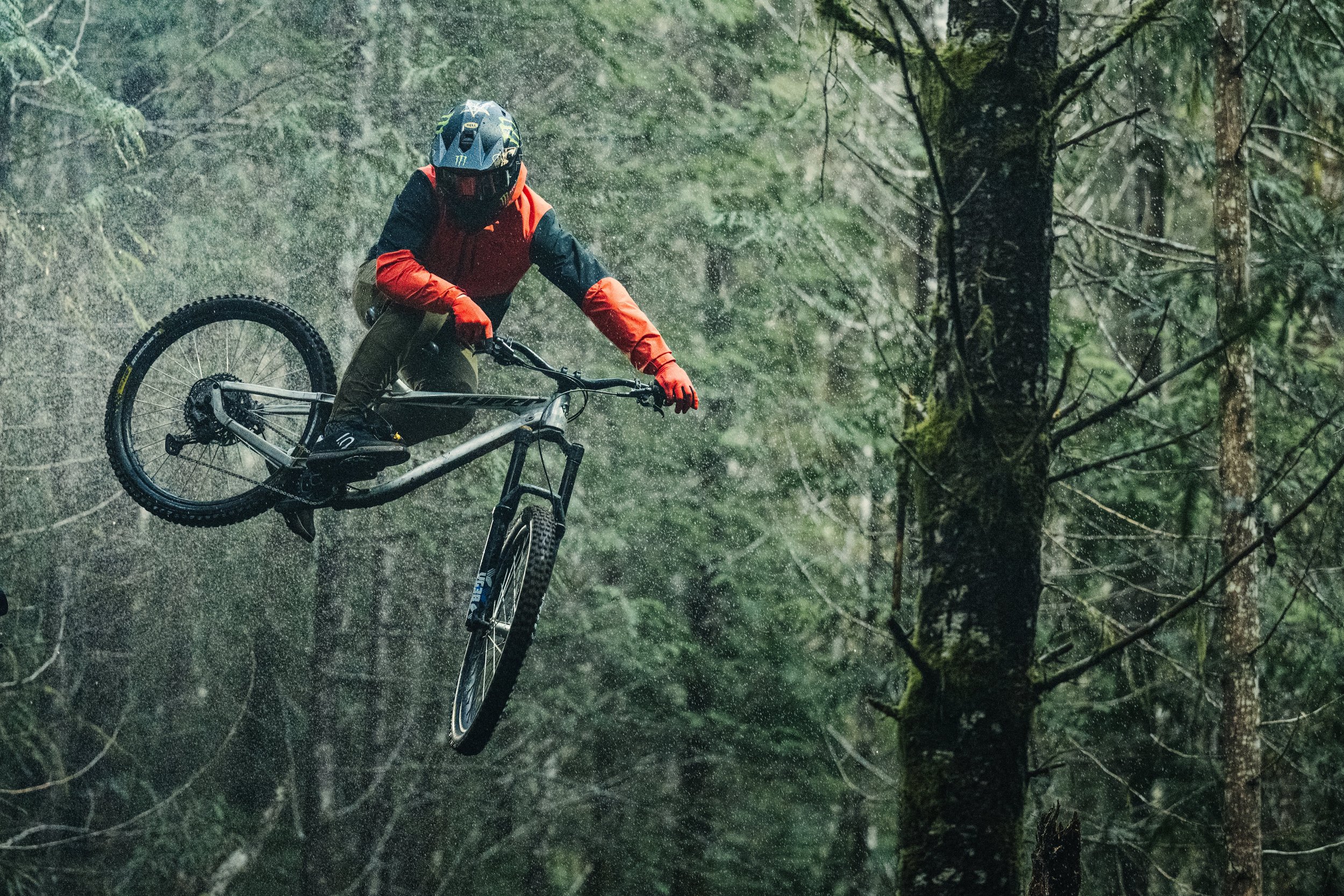 A person in a red and black jacket and helmet riding a mountain bike in mid-air above a forest trail, with trees and greenery in the background.